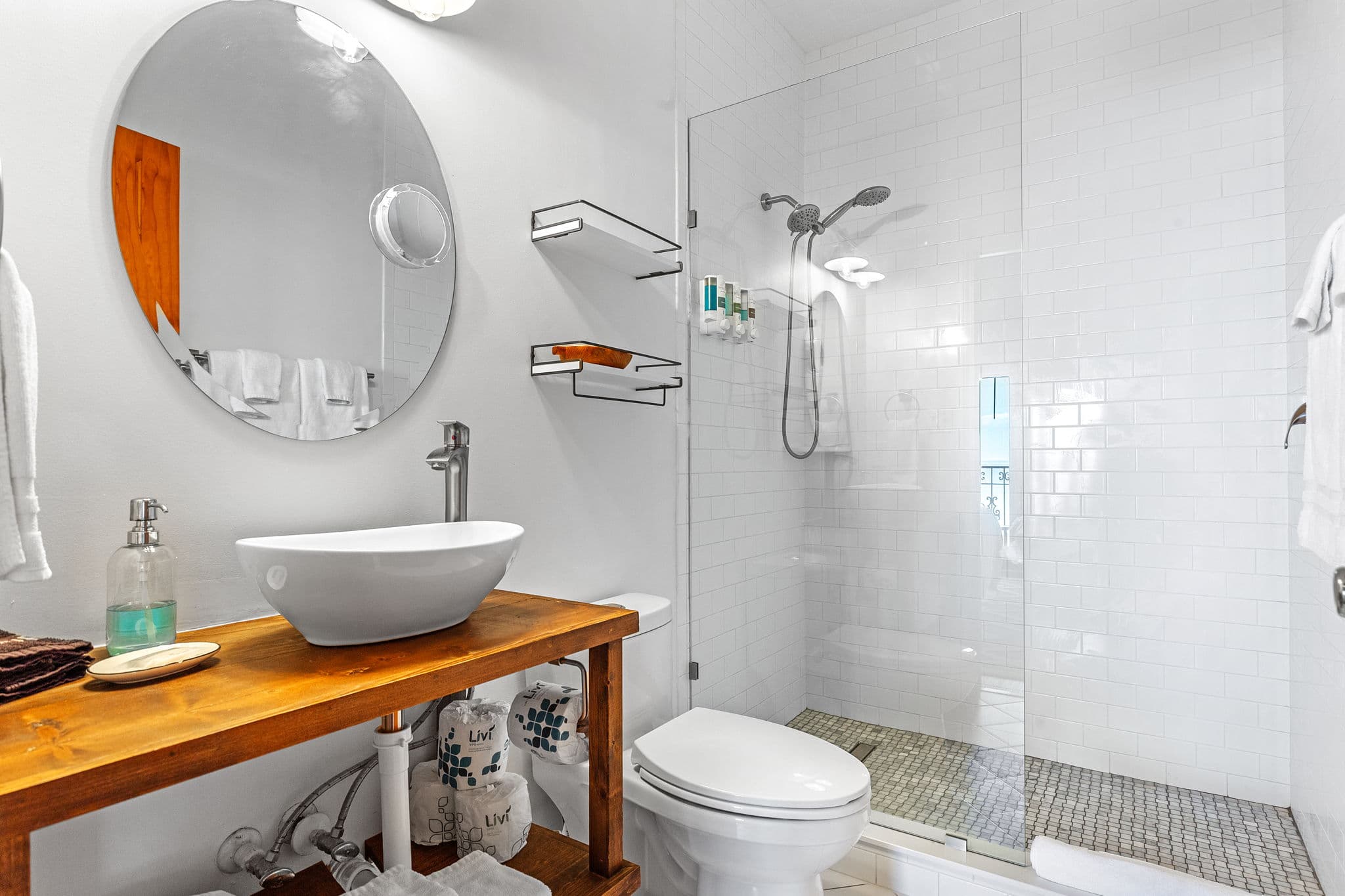 Sparkling clean white bathroom with tiled walk-in shower, toilet, wooden counter with white vessel sink with round frameless mirror mounted above