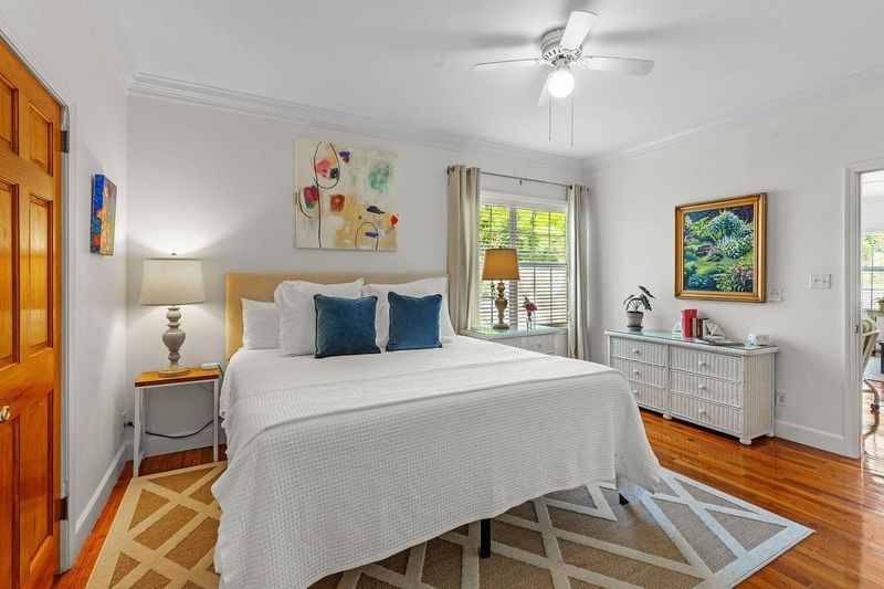 A bright and inviting bedroom with a king bed dressed in white linens and blue pillows. The room features hardwood floors, a ceiling fan, artwork on the walls, and a nightstand with a lamp.