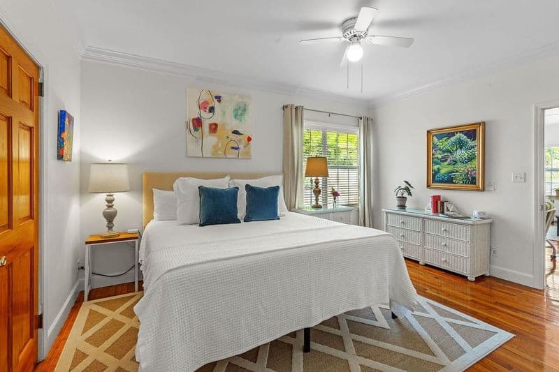 A bright and inviting bedroom with a king bed dressed in white linens and blue pillows. The room features hardwood floors, a ceiling fan, artwork on the walls, and a nightstand with a lamp.