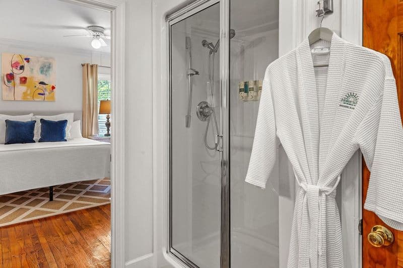 Private bathroom with shower on the right and a white bathrobe hanging on the door. In the background, a bedroom can be seen with a bed and artwork on the wall.
