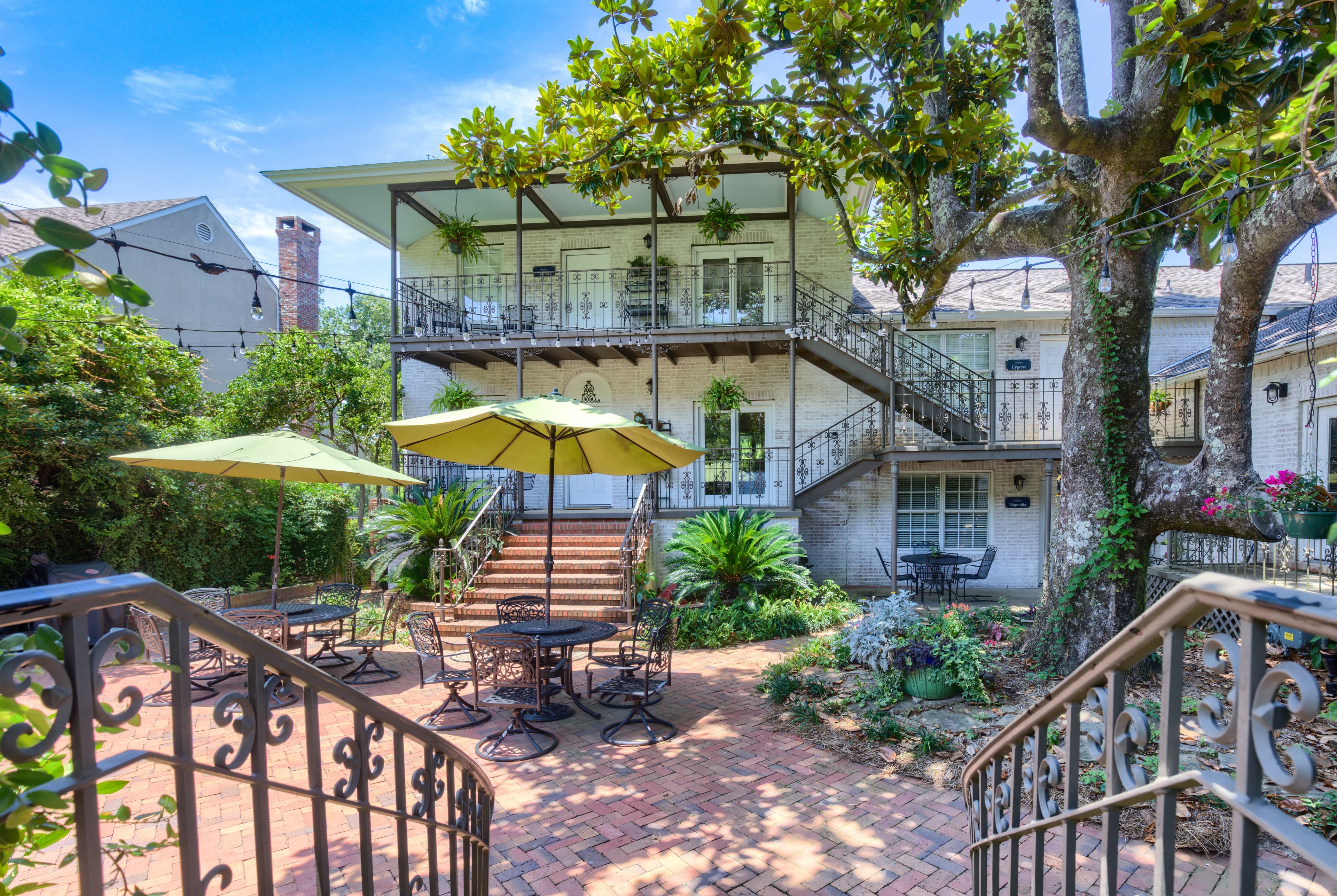 A charming courtyard with a brick walkway, lush greenery, and a two-story building with balconies. The courtyard is shaded by a large tree and features outdoor seating areas with tables and umbrellas.