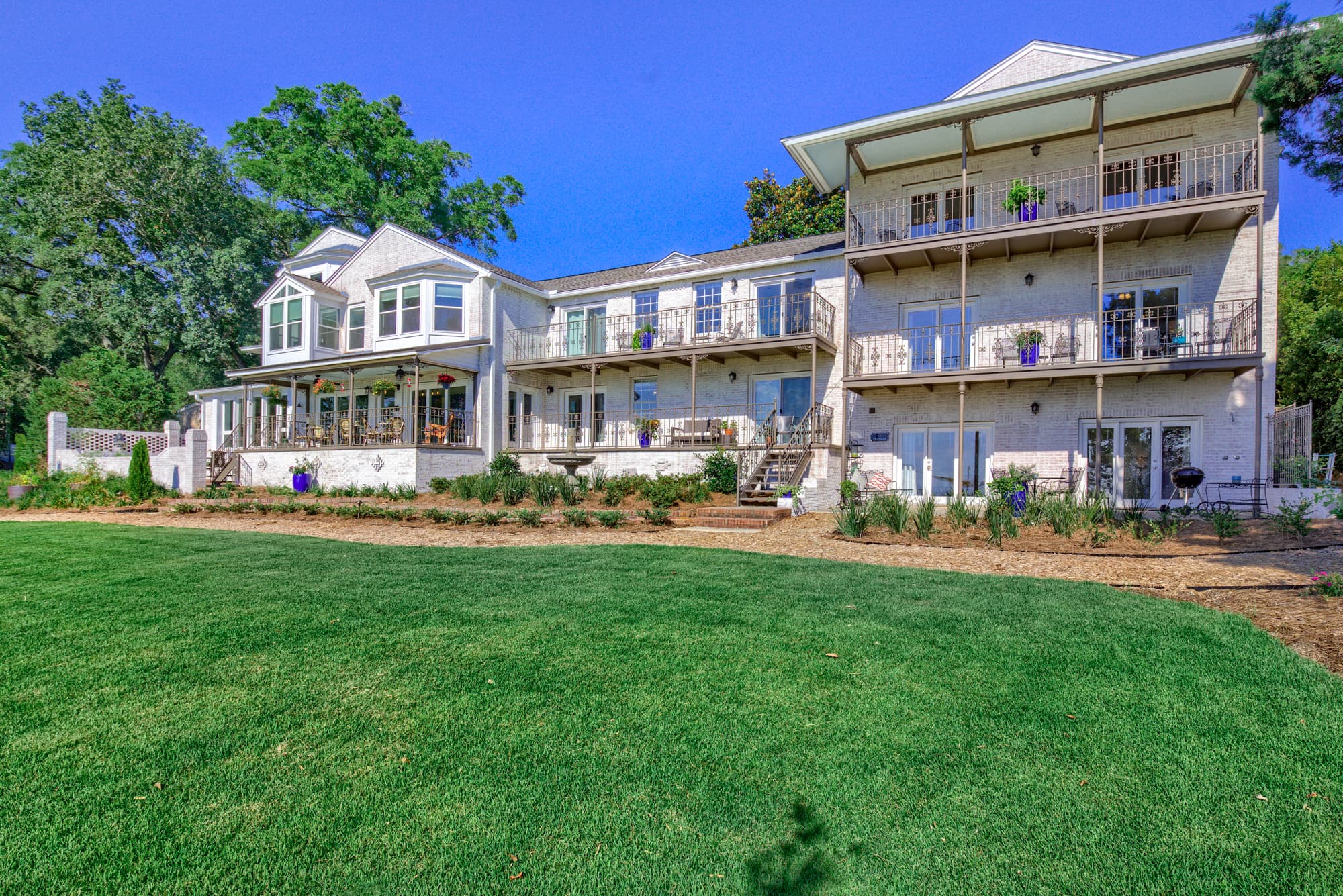 A large, white house with multiple levels and balconies overlooking a lush green lawn. The house has a classic architectural style with white columns and decorative details.