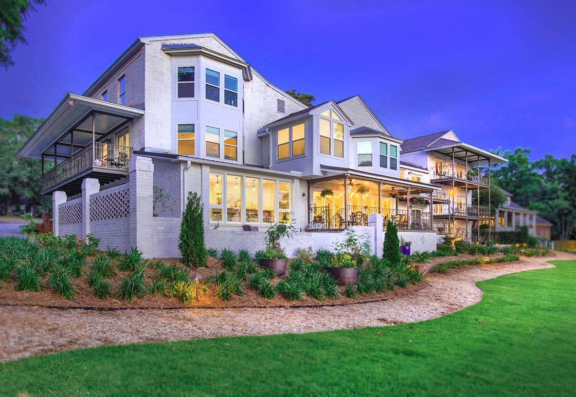 An expansive, two-story house with a white facade and multiple windows. The house features a wraparound porch and a landscaped backyard with a gravel path winding through lush greenery. The scene is illuminated by warm evening light.