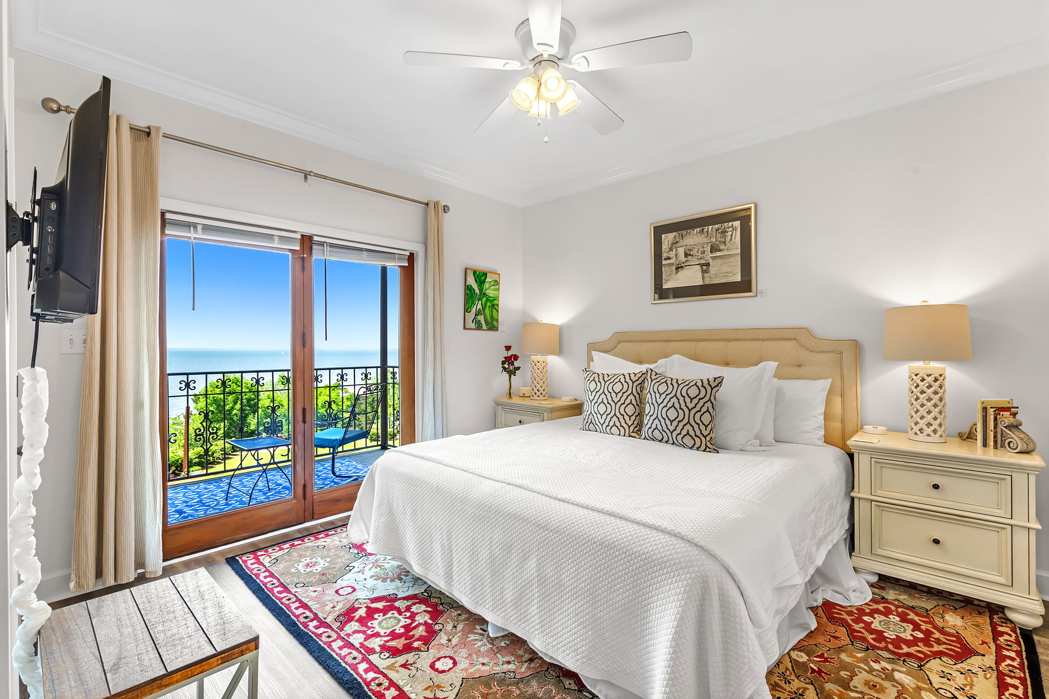 Bedroom with king bed topped with white bedding and taupe throw pillows, wooden nightstand with table lamp, red & gold oriental area rug, and sliding doors leading to balcony with bay views