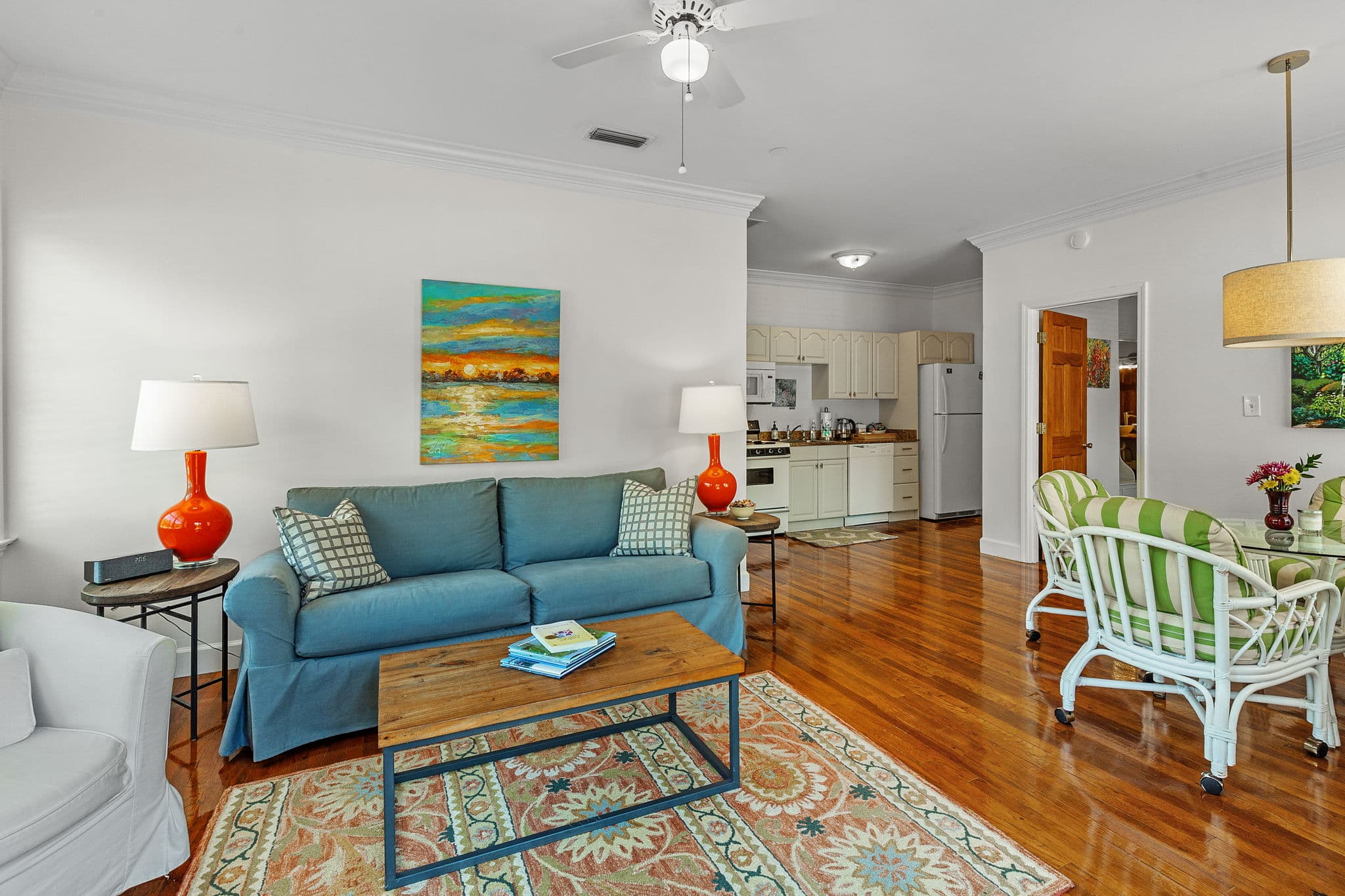 A cozy living room with a blue sofa, coffee table, and colorful rug. A dining area with a table and chairs is visible in the background. The room has hardwood floors and a ceiling fan.