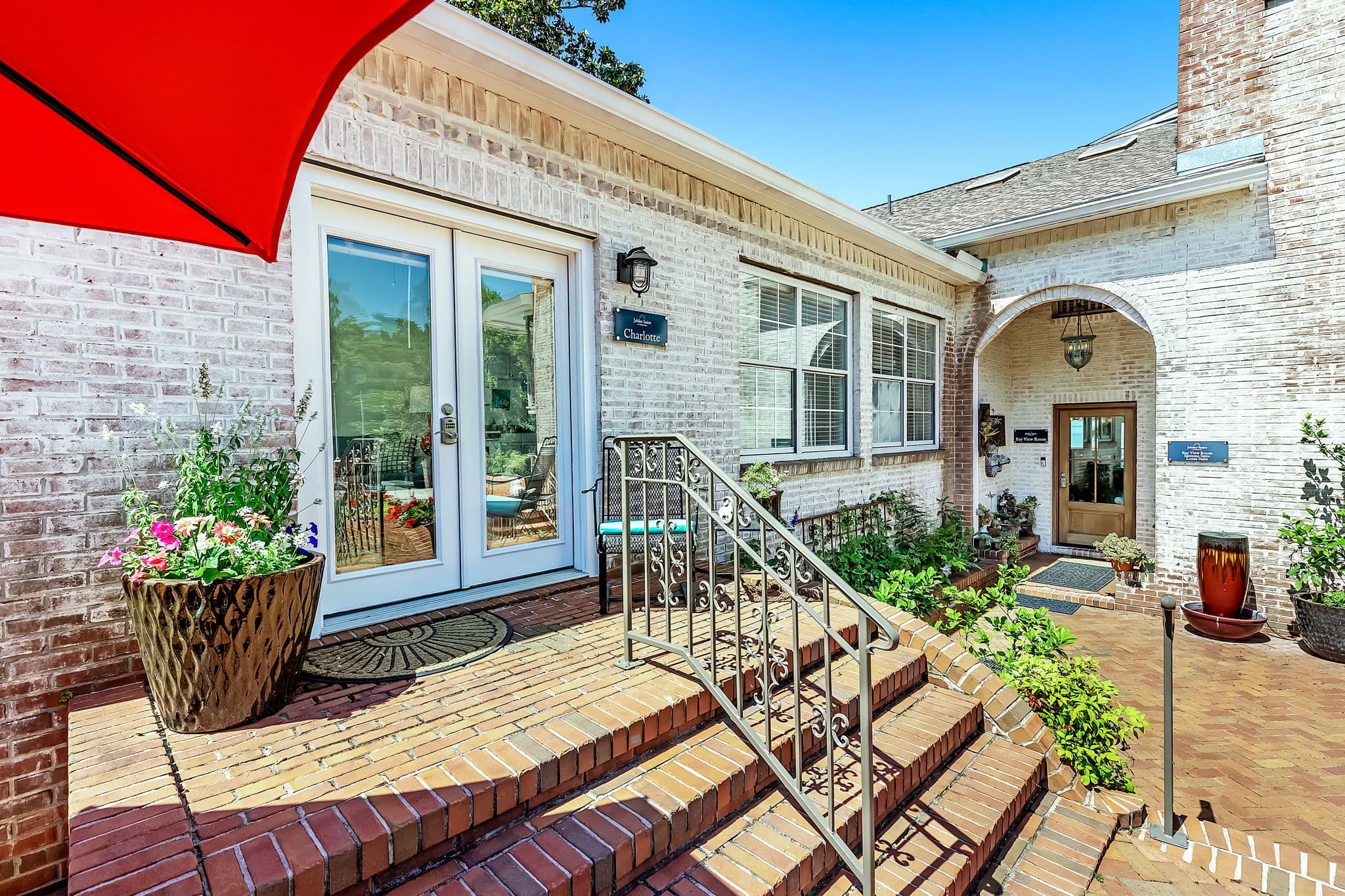 A charming outdoor entrance to a building with a red umbrella, brick steps, and potted plants. The building has white brick walls and multiple entrances.