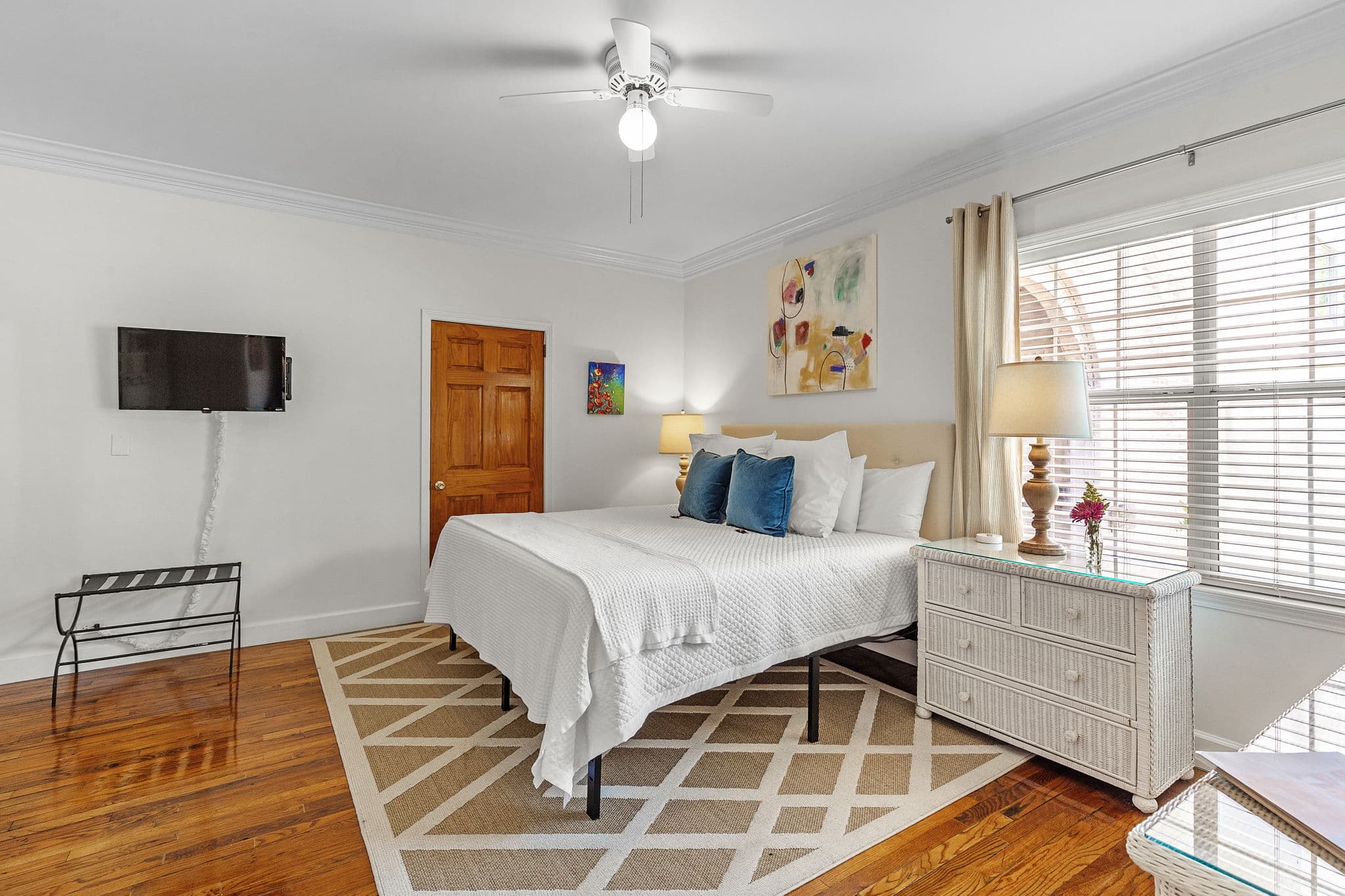 A cozy bedroom with a king bed, a TV mounted on the wall, and a patterned rug on the hardwood floor. The room features a large window that lets in natural light and a dresser with a lamp.