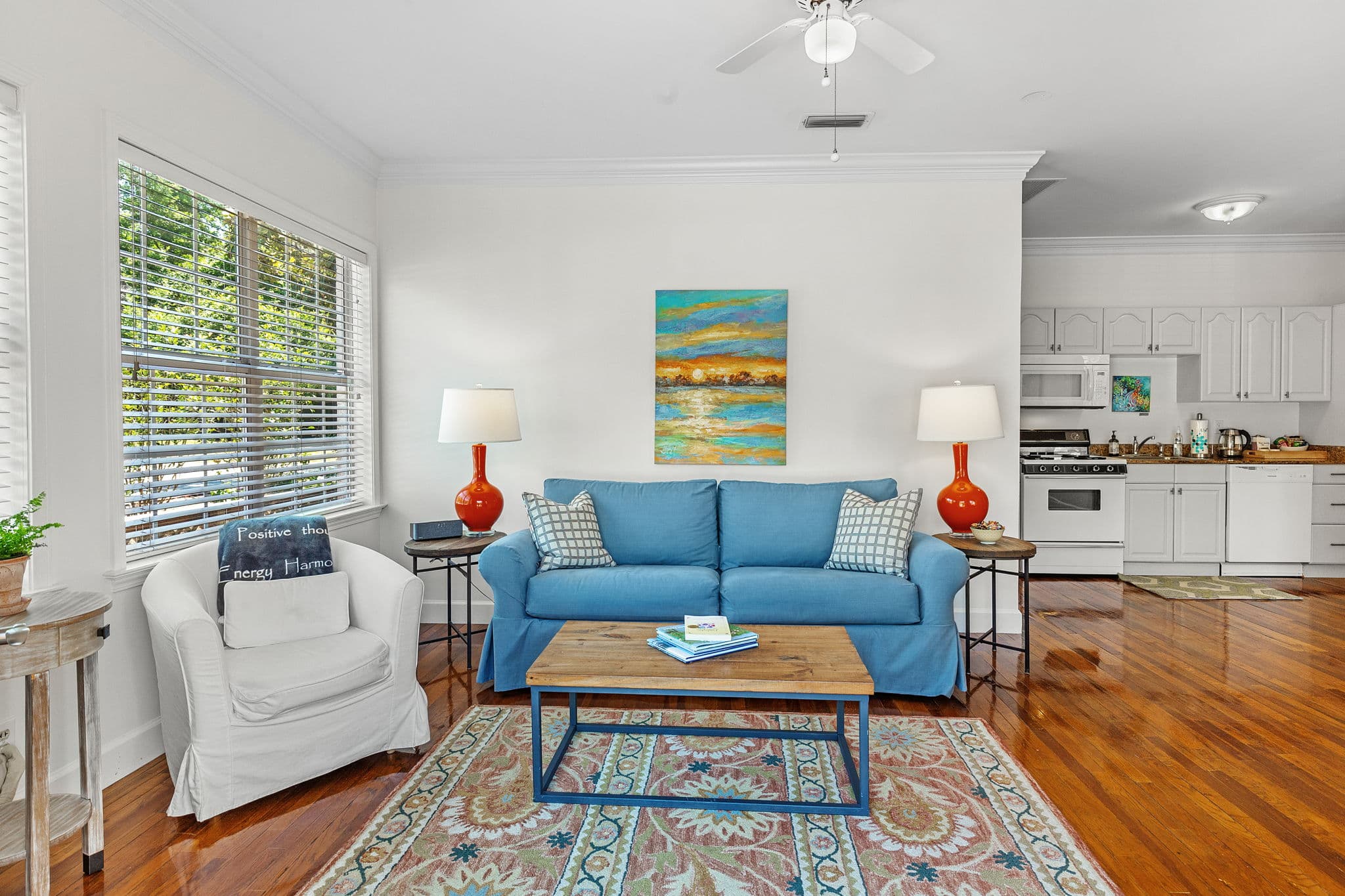 Main living space with a blue sofa, coffee table, and colorful rug. The full kitchen is visible in the background. The room has hardwood floors and a ceiling fan.
