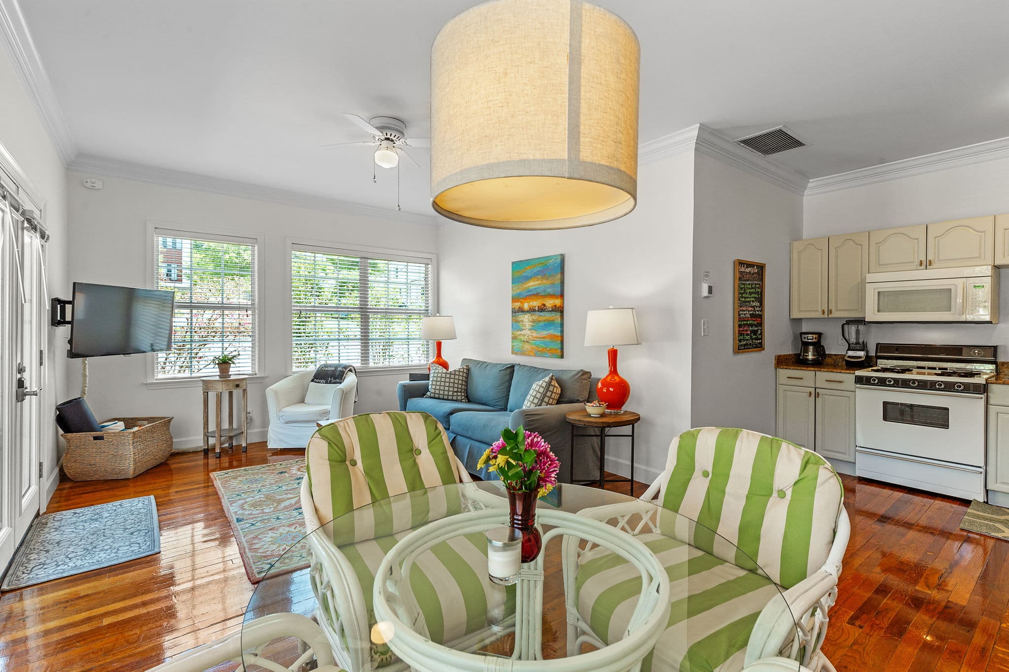 Seating area with a dining table and chairs in the foreground. A living room with a sofa and coffee table is visible in the background. The room has hardwood floors, a ceiling fan, and a large window that lets in natural light.