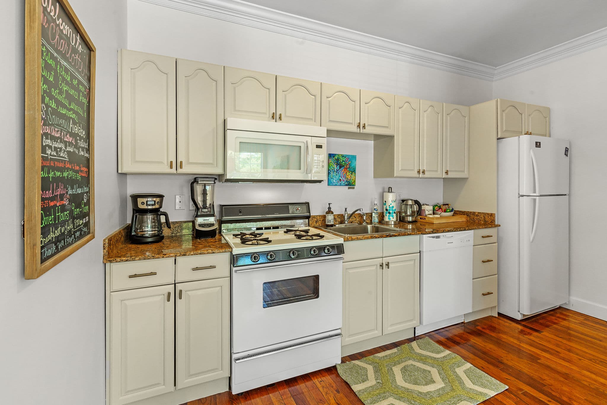 A well-equipped kitchen with white cabinets and appliances. The countertops are a warm wood tone, and there's a green rug on the hardwood floor.