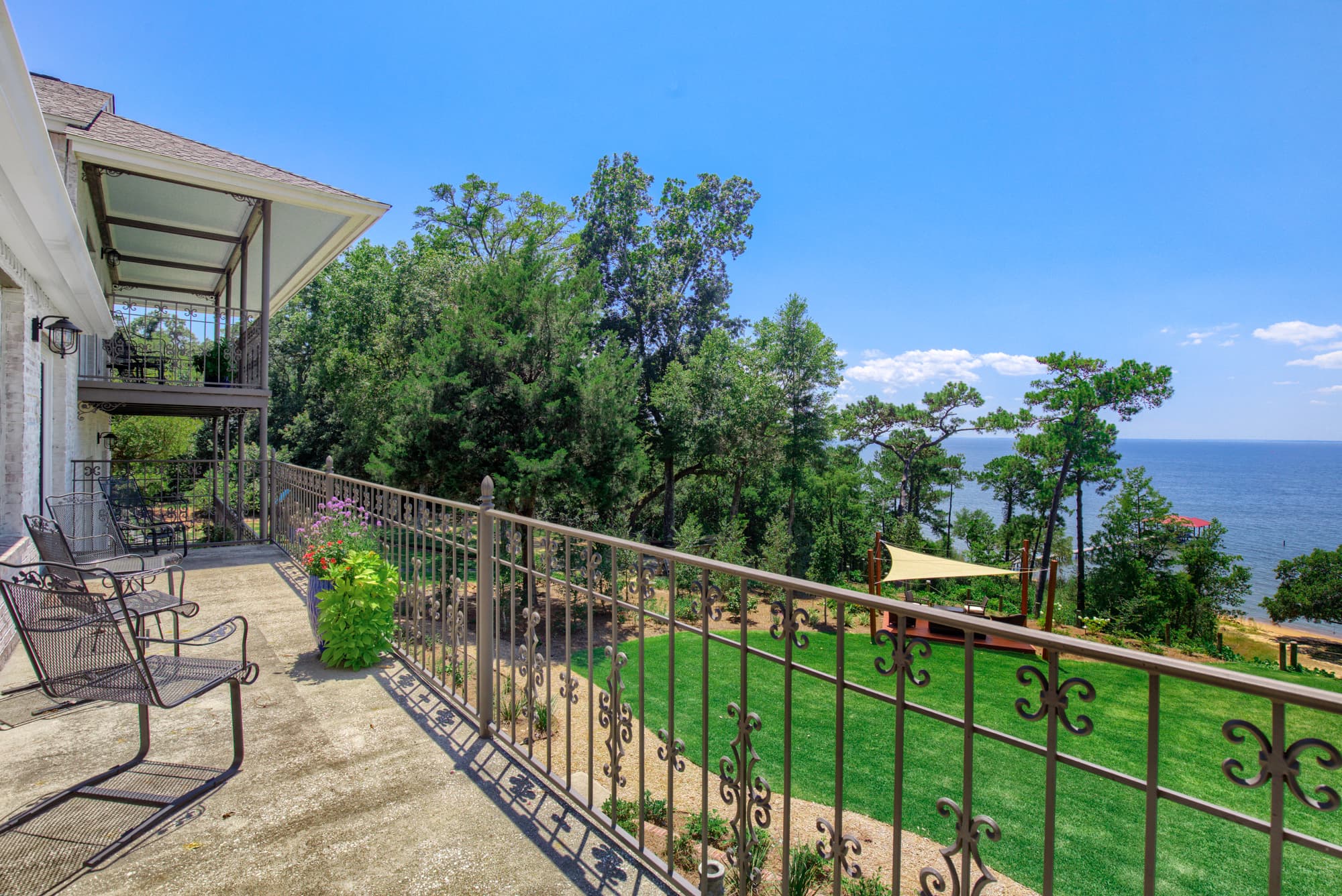 Balcony with metal chairs, decorative metal railing overlooking vibrant green lawn, trees, and gorgeous blue waters of the bay under a bright blue sky