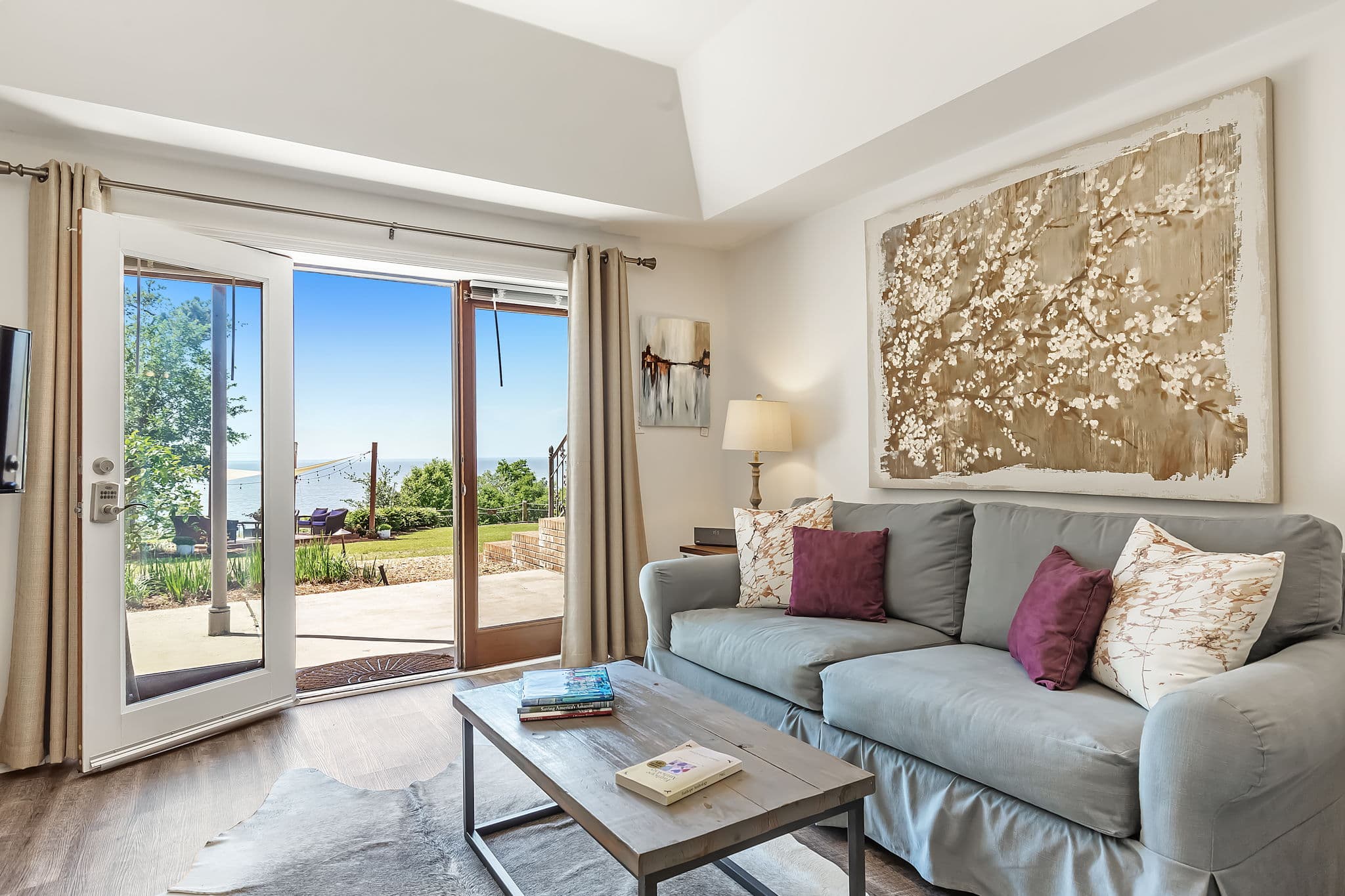 A bright, airy living room with a large gray sofa, a coffee table, and large artwork on the wall. Sliding glass doors open onto a patio with bay views.