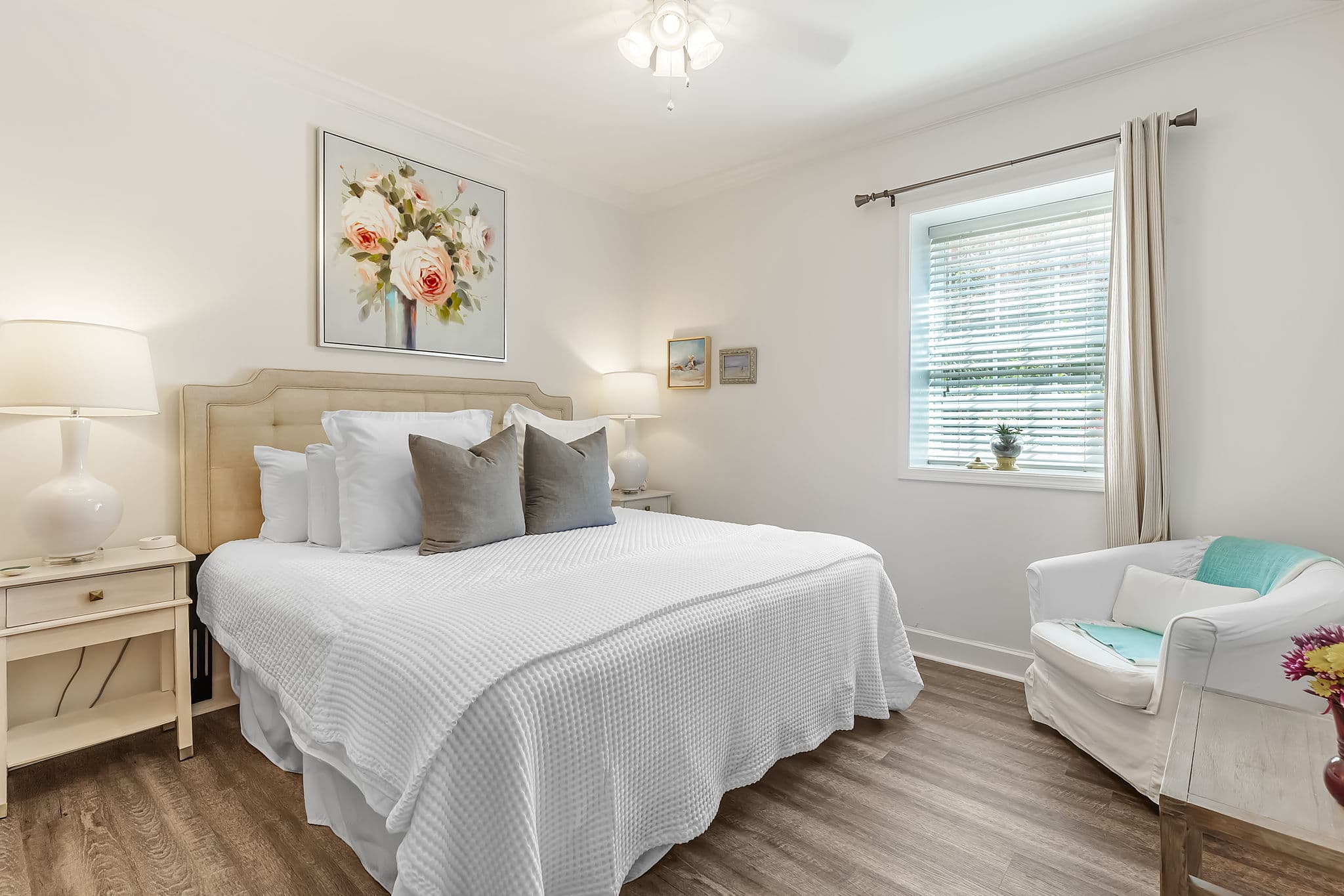 A light-filled bedroom with a king-size bed made with white linens and gray throw pillows. There's a white wooden headboard and a matching nightstand with a lamp. A white armchair with a blue throw sits in the corner, and a large floral painting hangs on the wall.