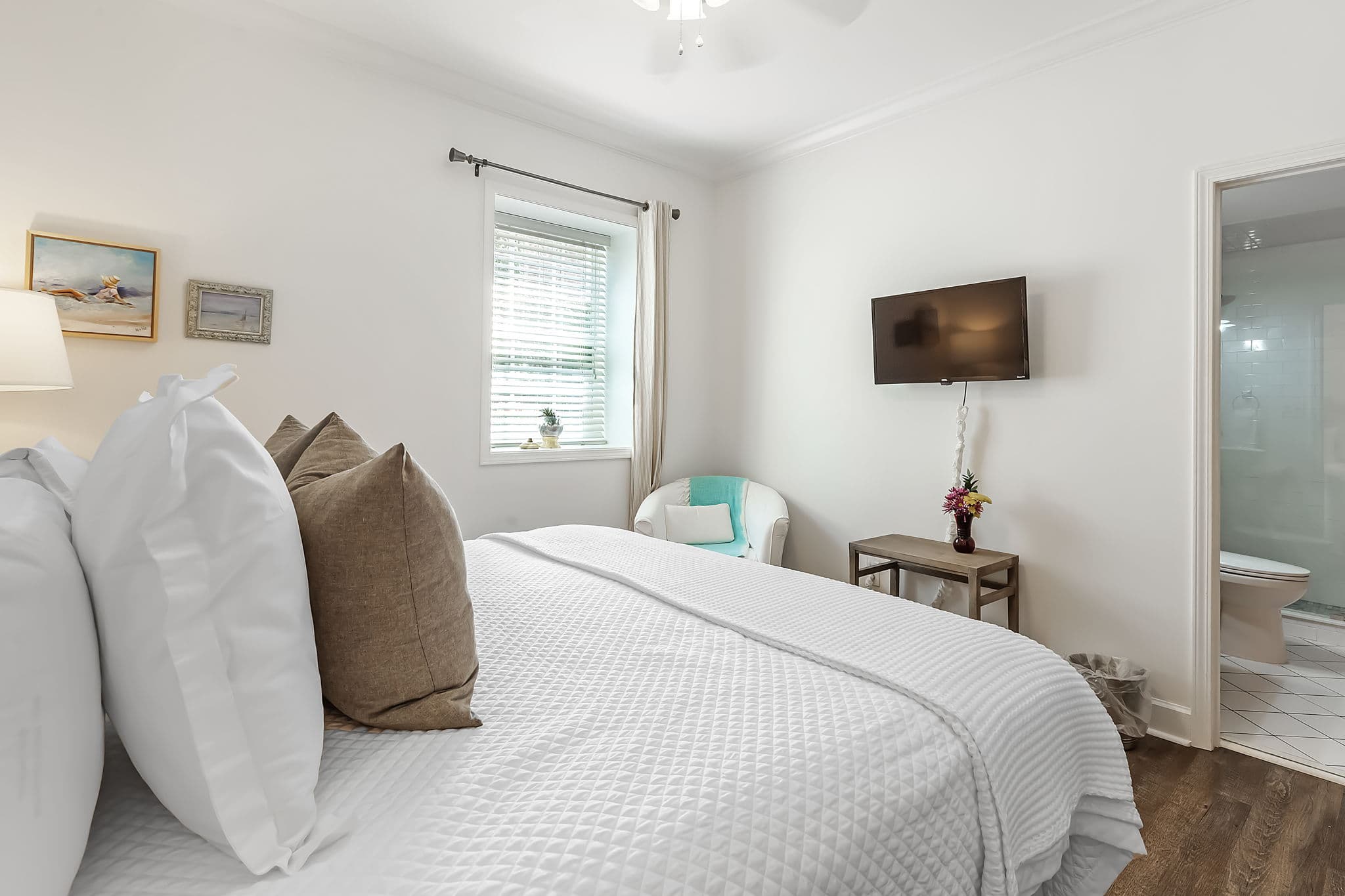 A light and airy bedroom with a king-size bed made with white linens and throw pillows. A flat-screen TV is mounted on the wall, and a white armchair with a blue throw sits in the corner.