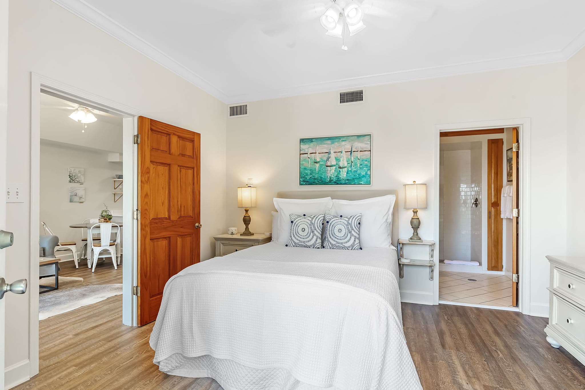 A bright and airy bedroom with a queen-size bed made with white linens and blue throw pillows. There's a white wooden headboard and matching nightstands with lamps. An open doorway reveals a living area with a dining table and chairs.