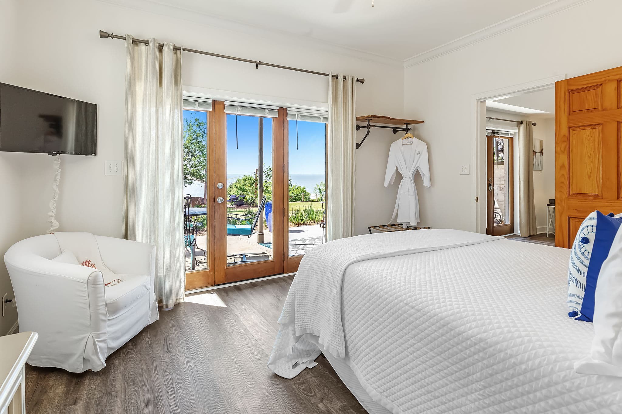 An inviting bedroom with a queen-size bed made with white linens and blue throw pillows. Sliding glass doors lead to a patio with bay views. A white armchair sits in the corner, and a flat-screen TV hangs on the wall.