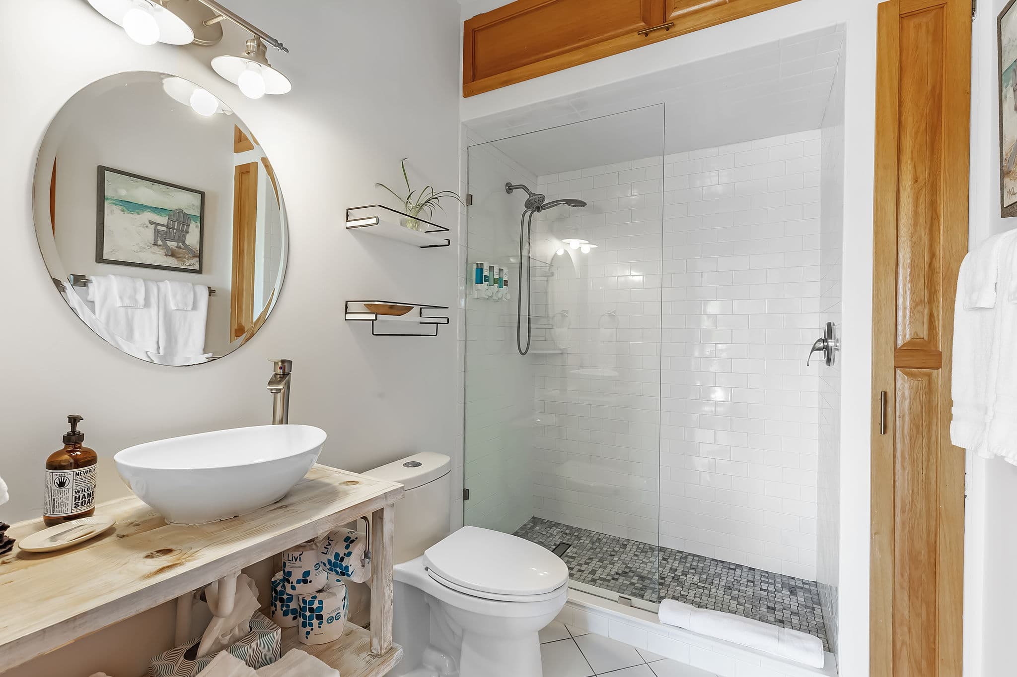 A clean and inviting bathroom with a white tiled shower and a round mirror above a wooden vanity. The vanity has a vessel sink and storage space below. A white toilet sits next to the shower.