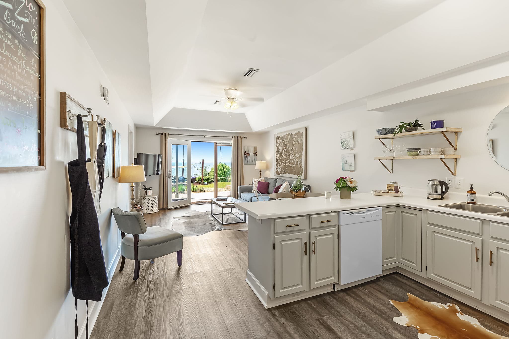 A bright and open living space with a kitchen, dining area, and living room. The kitchen has white cabinets and countertops. The living room has a gray sofa, a coffee table, and a sliding glass door leading to a patio with a view.