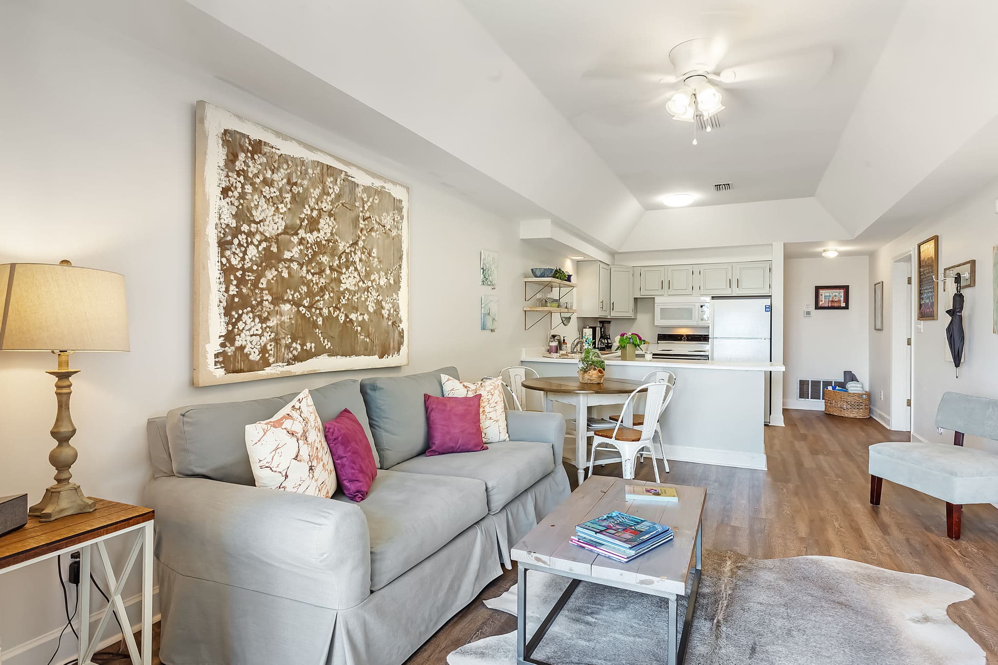 A comfy living room with a gray sofa, a coffee table, and large artwork on the wall. The kitchen is visible in the background with white cabinets and a dining table.
