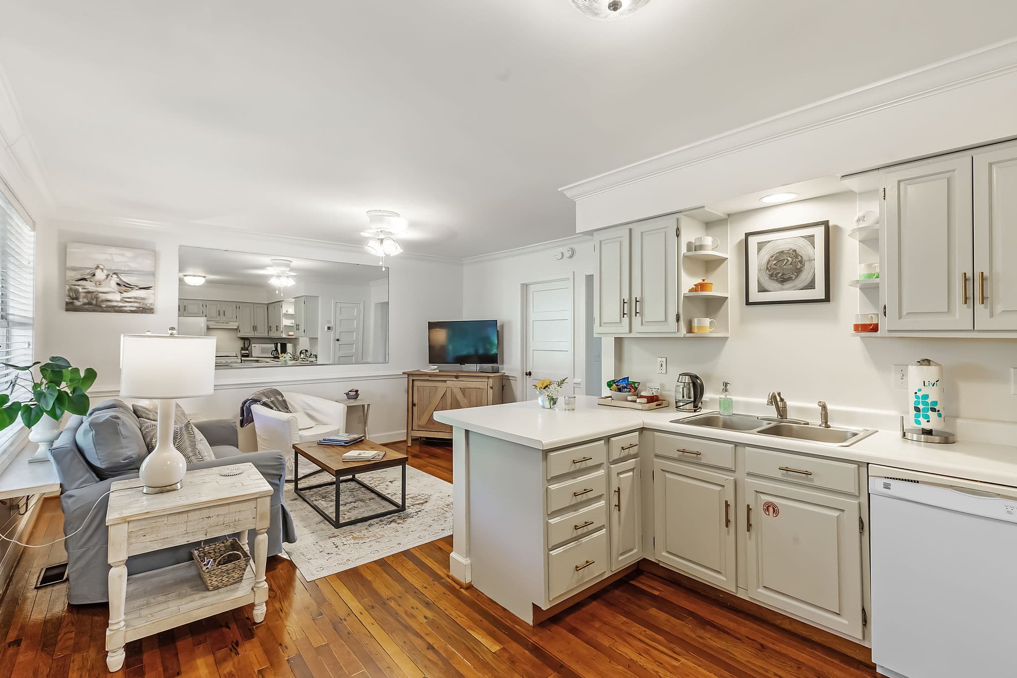 A bright and open kitchen and living room area. The kitchen features white cabinets, a sink, and a countertop. The living room has a comfortable sofa, a coffee table, and a TV. The space has hardwood floors and a bright, airy feel.