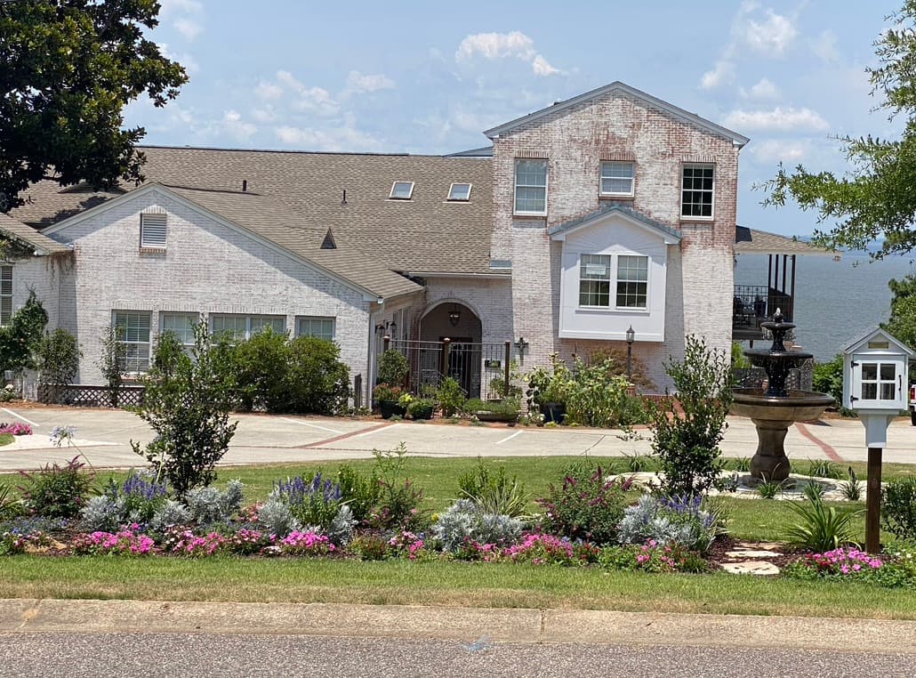 A large house with a white brick exterior and a fountain in the front yard. The house has a multi-level design with multiple windows and a large driveway. There are flower beds along the edge of the property.