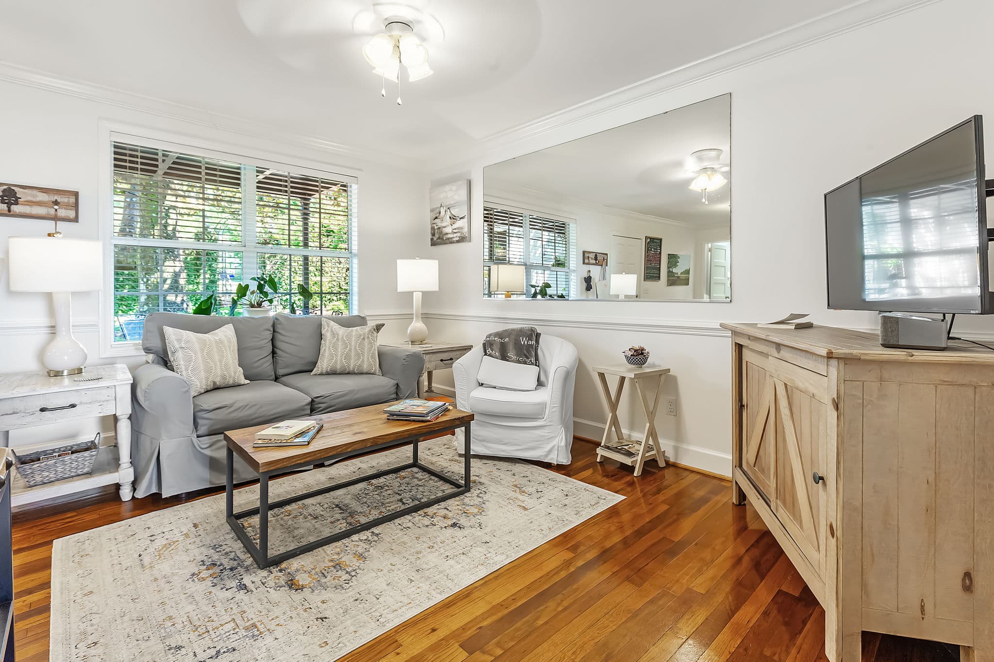 Main living area with a gray sofa, coffee table, and a colorful rug. A large mirror reflects the room and a TV is mounted on the wall. The room has hardwood floors and a ceiling fan.