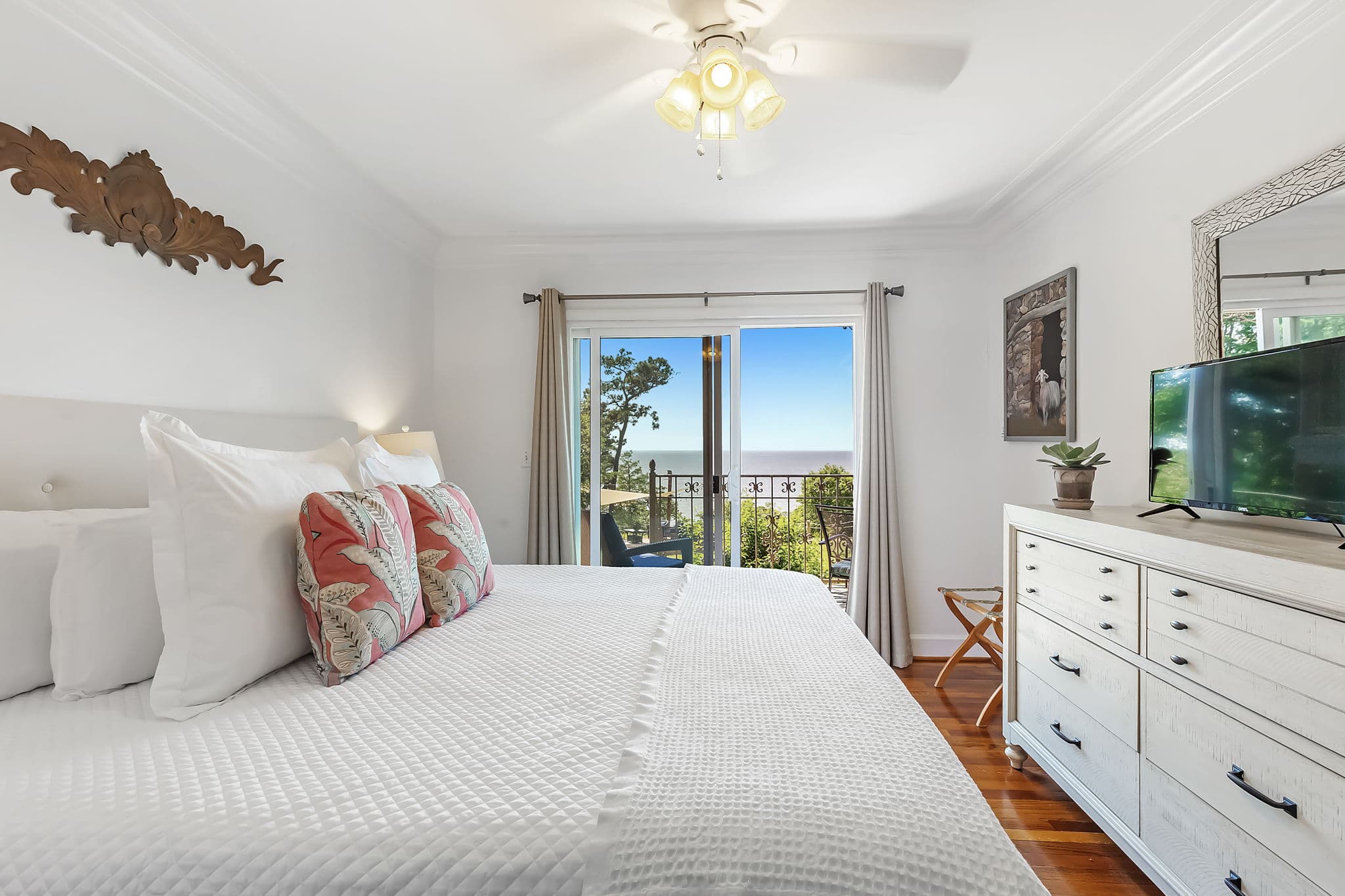 A bright and inviting bedroom with a king bed dressed in white linens and colorful pillows. The room features a ceiling fan, a TV mounted on the wall, a dresser, and sliding glass doors that open to a balcony with a view of the water..