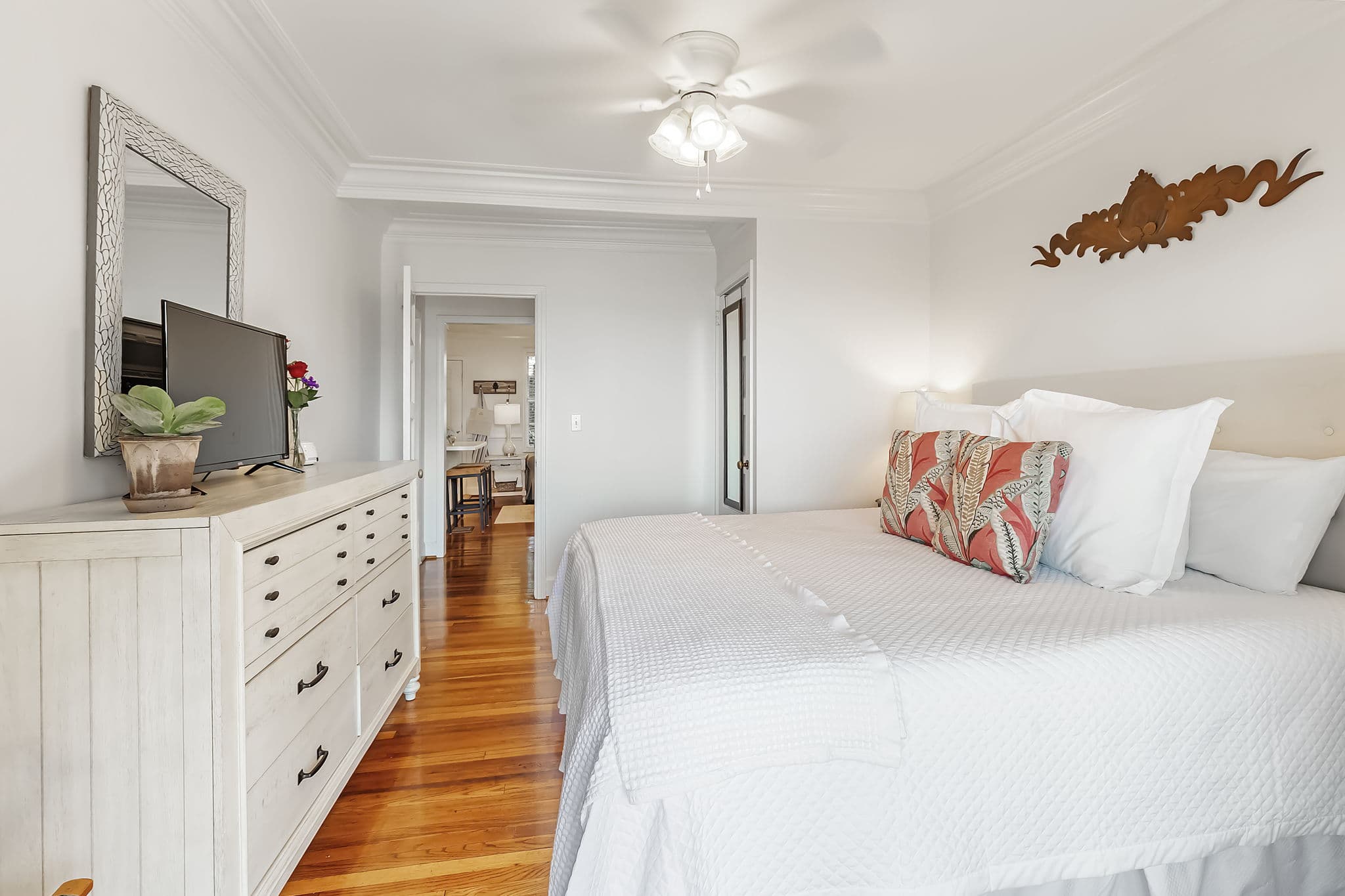A cozy bedroom with a king bed dressed in white linens and colorful pillows. The room features a ceiling fan, a TV mounted on the wall, and a dresser. Hardwood floors and a mirror add to the warm and inviting atmosphere.