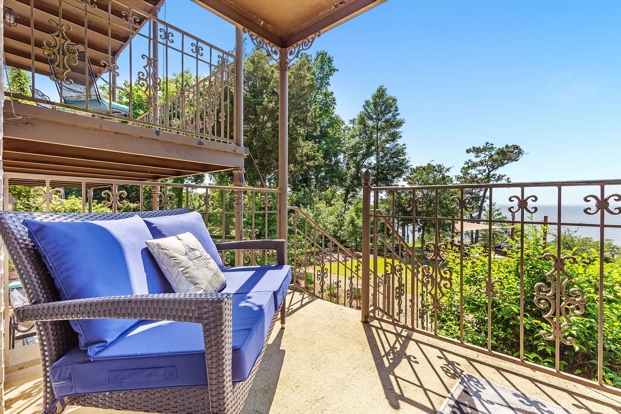A cozy outdoor seating area with a blue cushioned bench overlooking a scenic vista. The view includes lush greenery and a glimpse of the ocean in the distance.