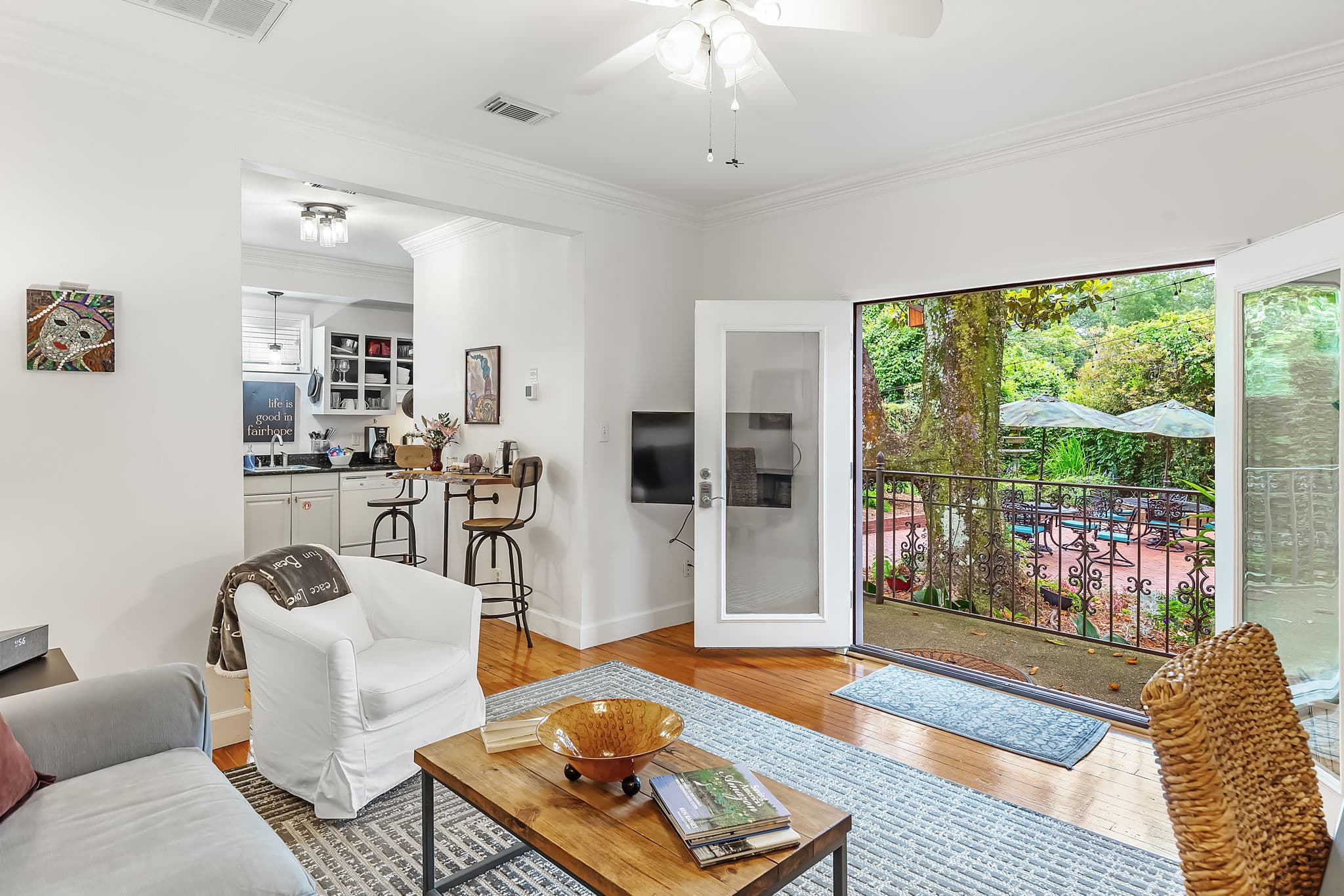 Sitting area of the New Orleans room features a comfortable sofa and armchair, a coffee table, and a colorful rug. The room has hardwood floors, a ceiling fan, and a glimpse of the kitchen can be seen in the background. Large glass doors open onto courtyard.