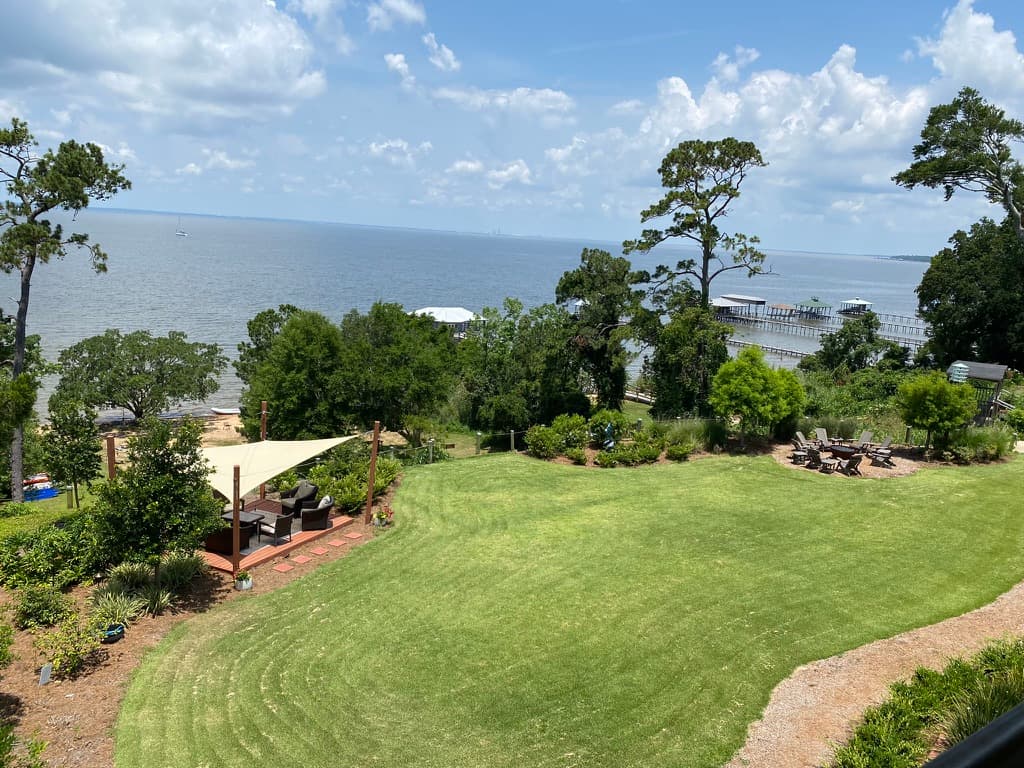 A scenic view from the bluff. A vast expanse of green lawn stretches towards the water's edge. In the distance, a calm body of water stretches out under a bright blue sky dotted with fluffy white clouds. Trees frame the edges of the view, adding depth and perspective.