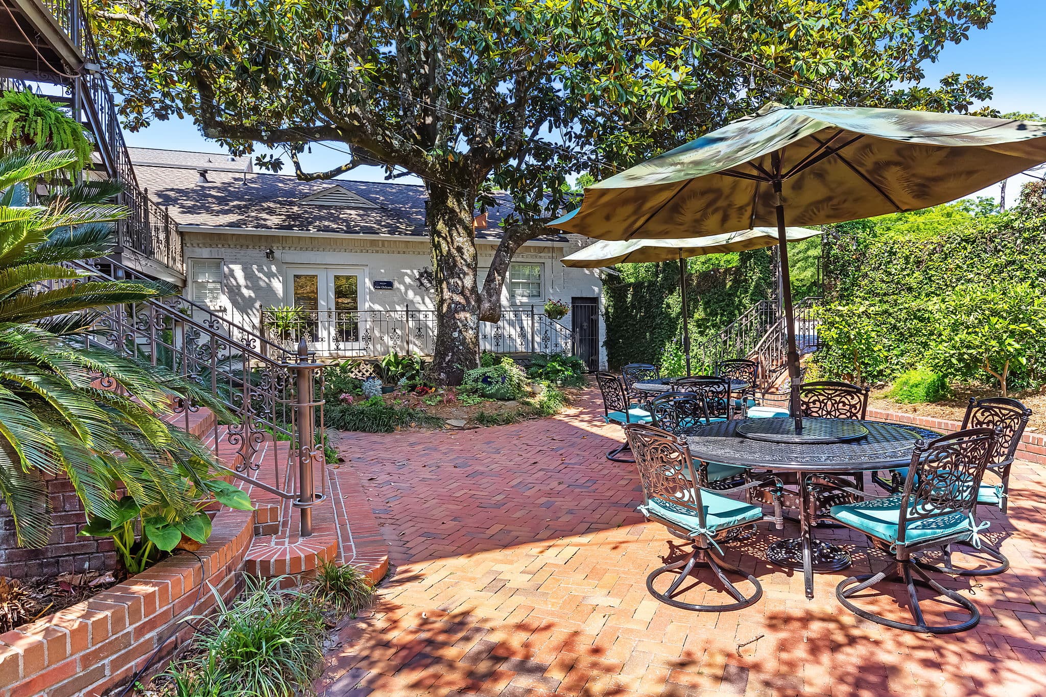 A charming courtyard with a brick walkway and lush greenery. A large tree provides shade, and there are several tables and chairs set up for outdoor dining. Umbrellas offer additional shade and protection from the sun.