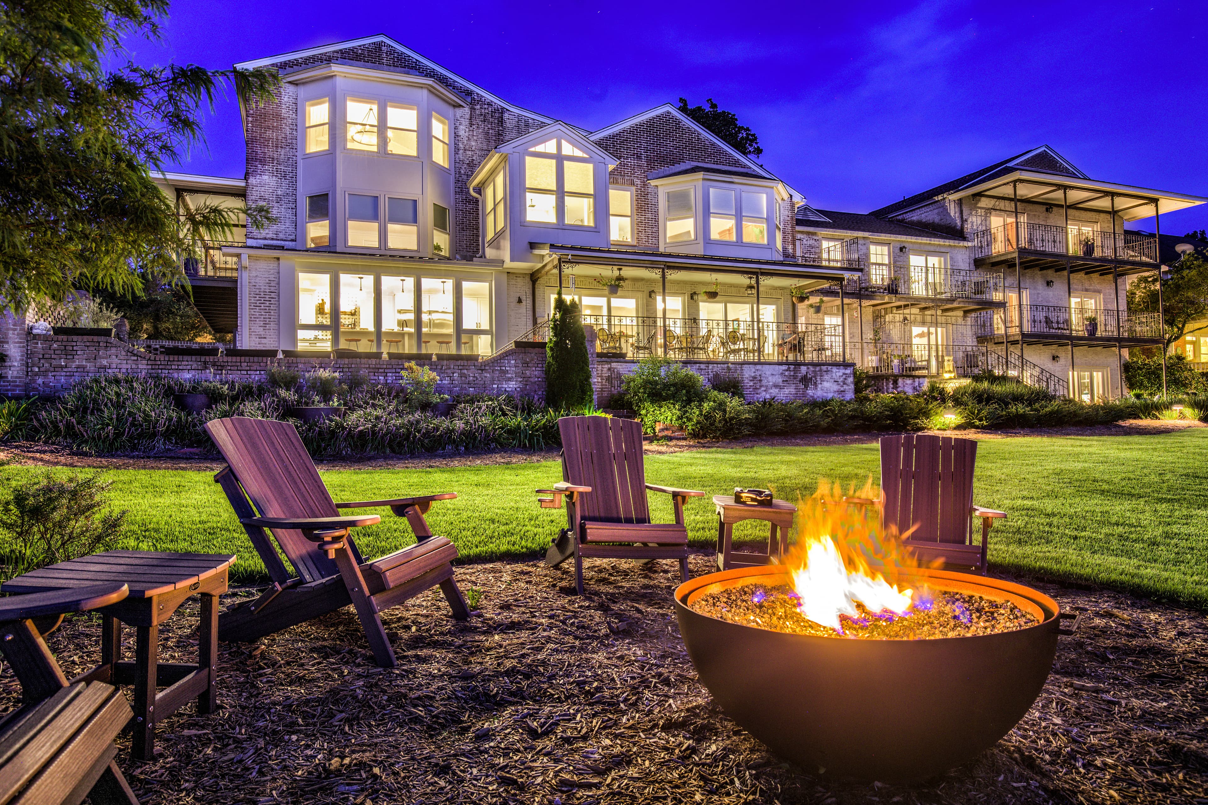 A cozy outdoor scene at dusk. Jubilee Suites glows with warm lighting illuminating the background. In the foreground, a fire pit crackles with flames, surrounded by comfortable chairs perfect for gathering around. The scene evokes a sense of warmth and relaxation.