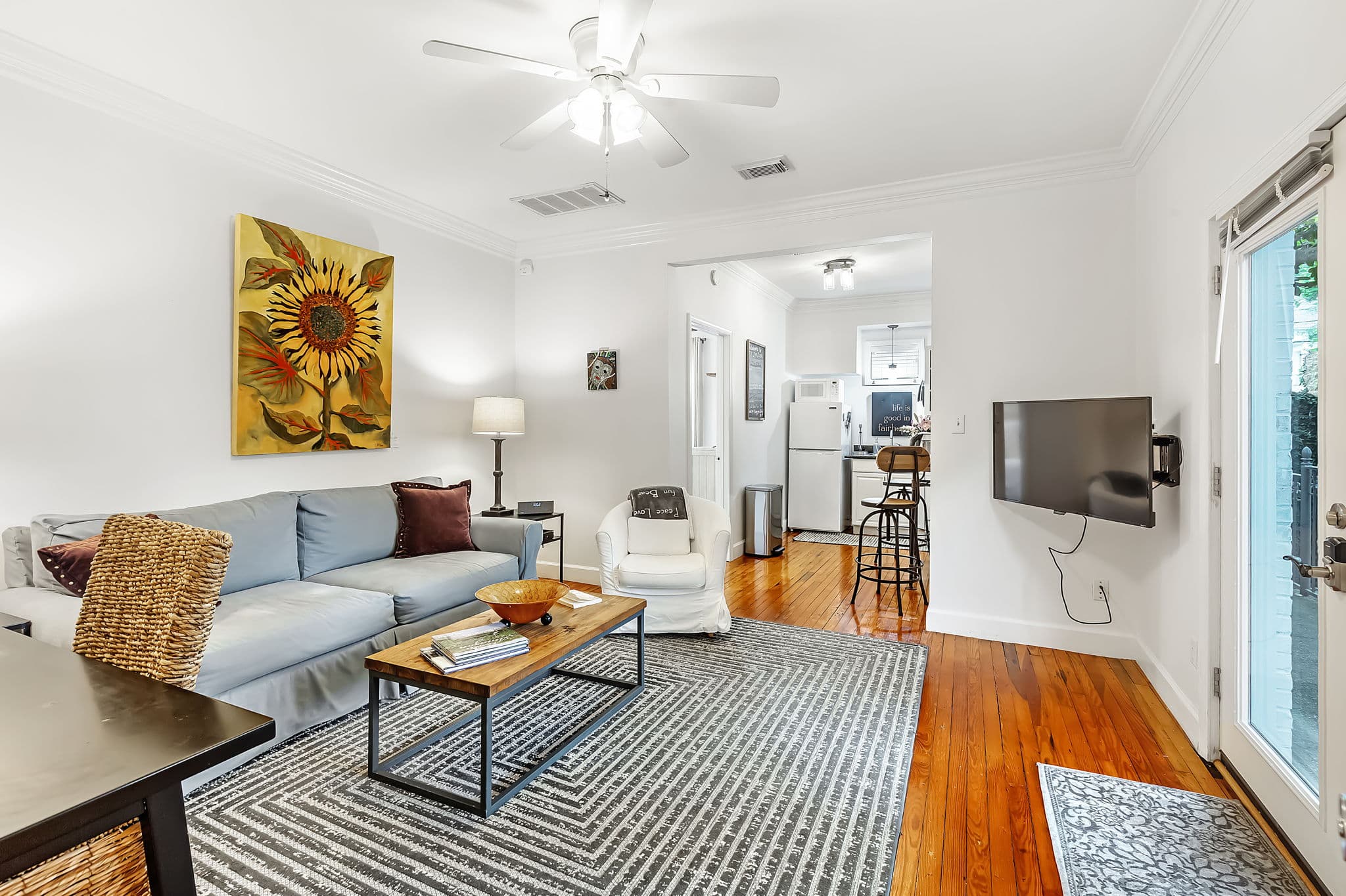Main living area with a blue sofa, coffee table, and colorful rug. A dining area with a table and chairs is visible in the background. The room has hardwood floors, a ceiling fan, and a TV mounted on the wall.
