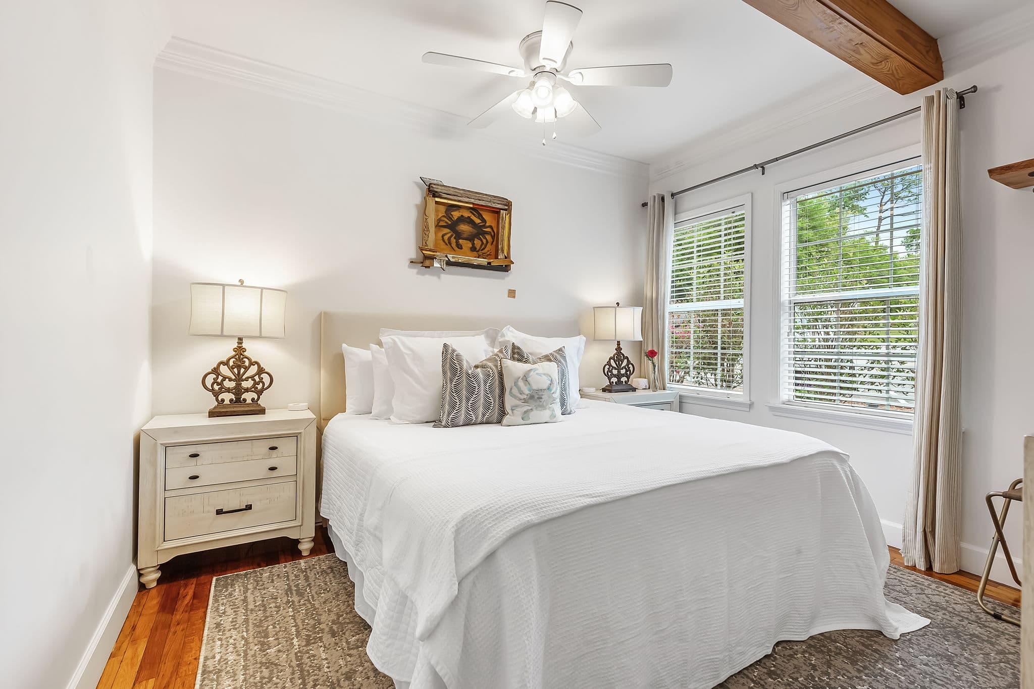A cozy bedroom with a king bed dressed in white linens and blue pillows. The room features hardwood floors, a ceiling fan, artwork on the walls, and a nightstand with a lamp.