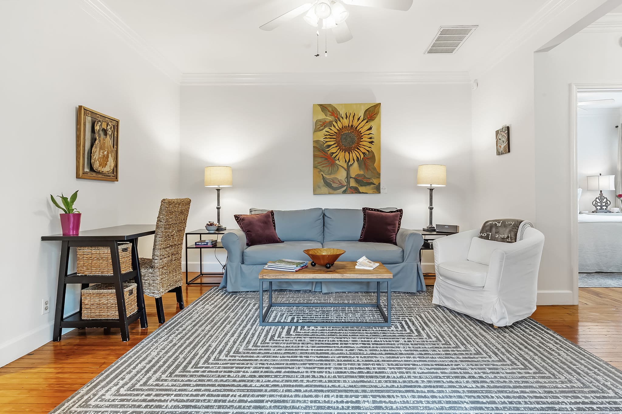 Living area with a blue sofa, coffee table, and colorful rug. A dining area with a table and chairs is visible in the background. The room has hardwood floors and a ceiling fan.