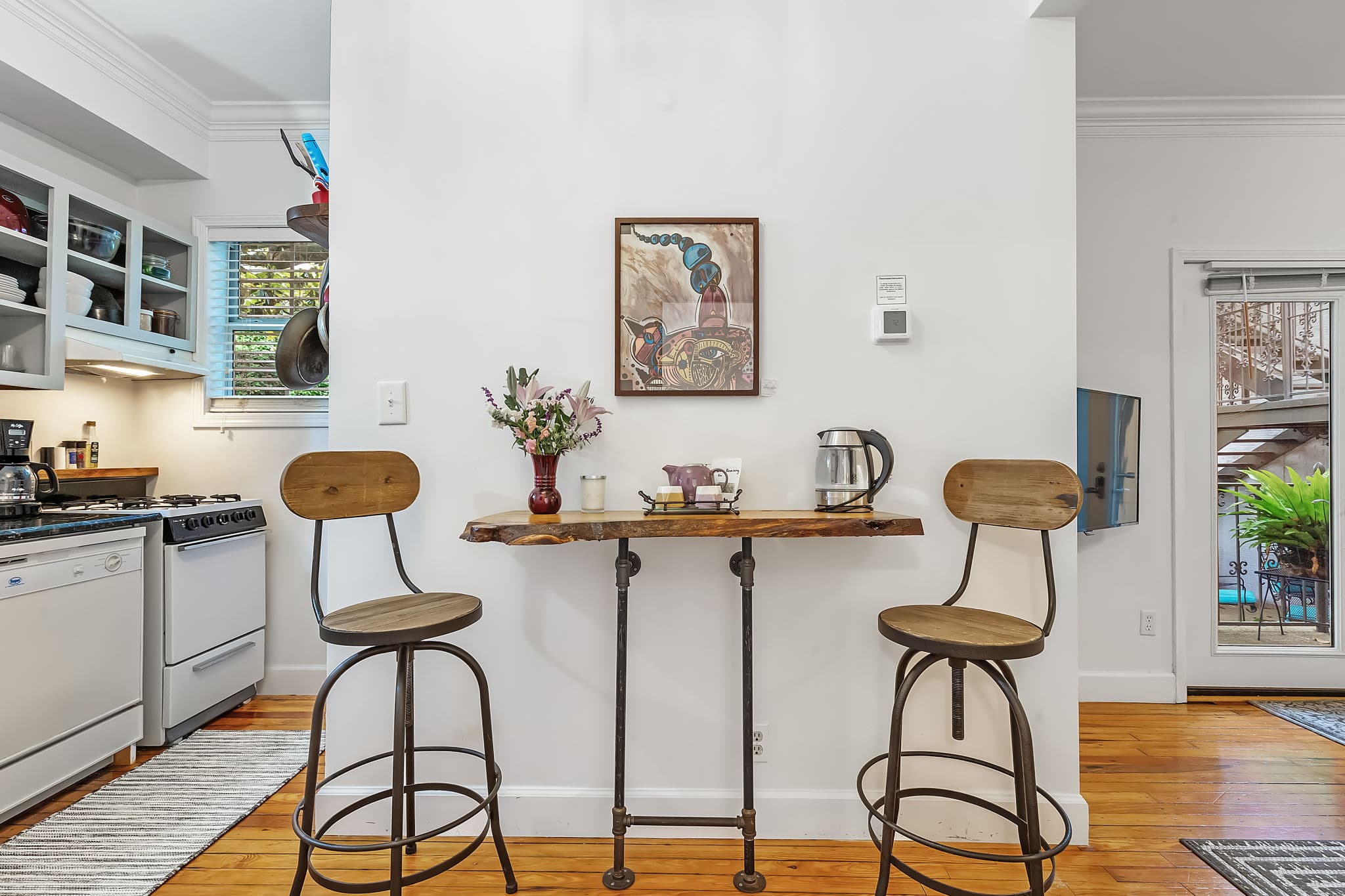 A cozy breakfast nook with a small table and two stools. The table has a rustic wooden top and metal legs, and the stools have a similar industrial style. There is a vase of flowers on the table and a framed picture on the wall.