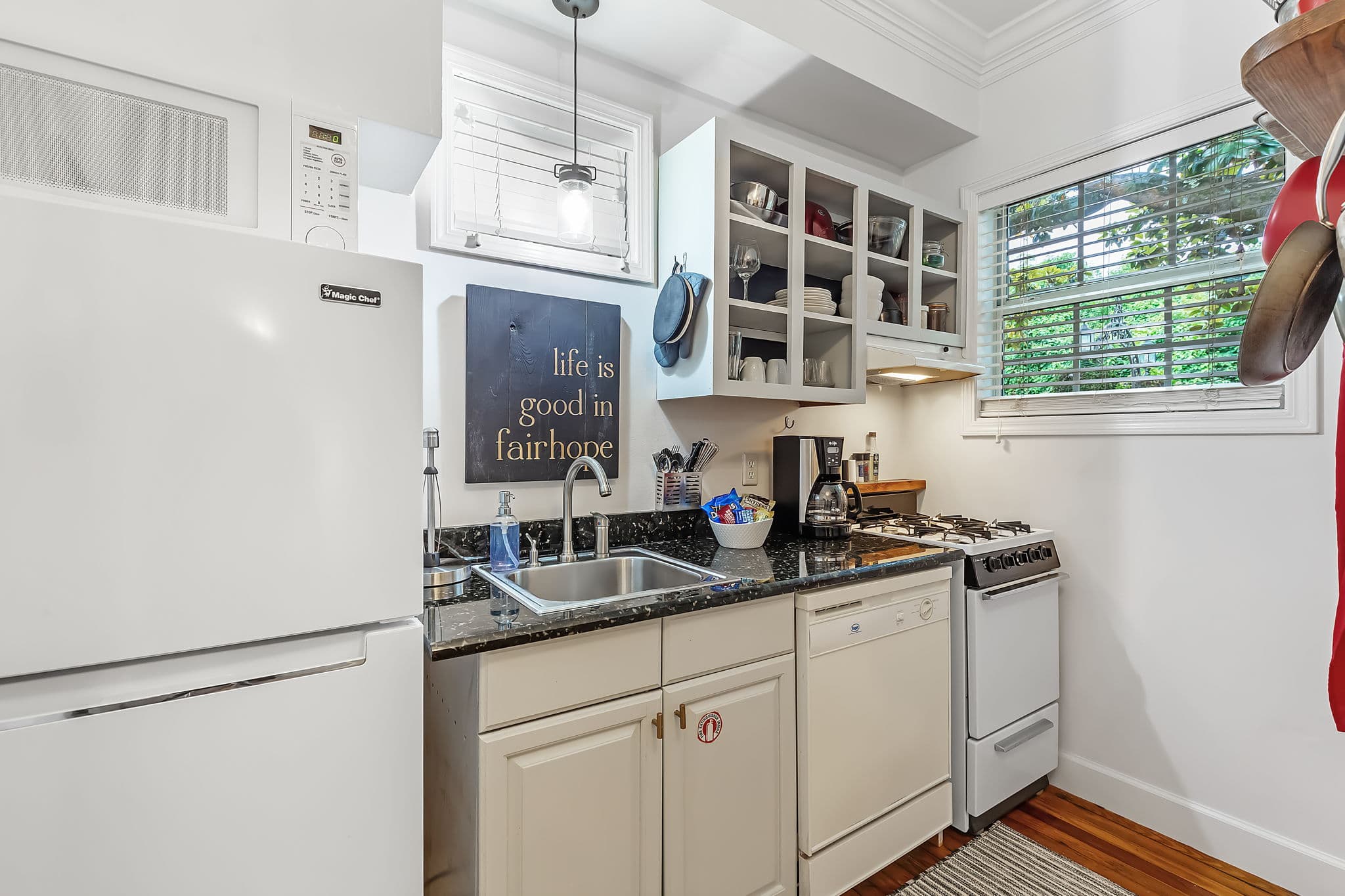 A compact but well-equipped kitchen with white cabinets and appliances. The countertops are black, and there is a window overlooking a lush green area.