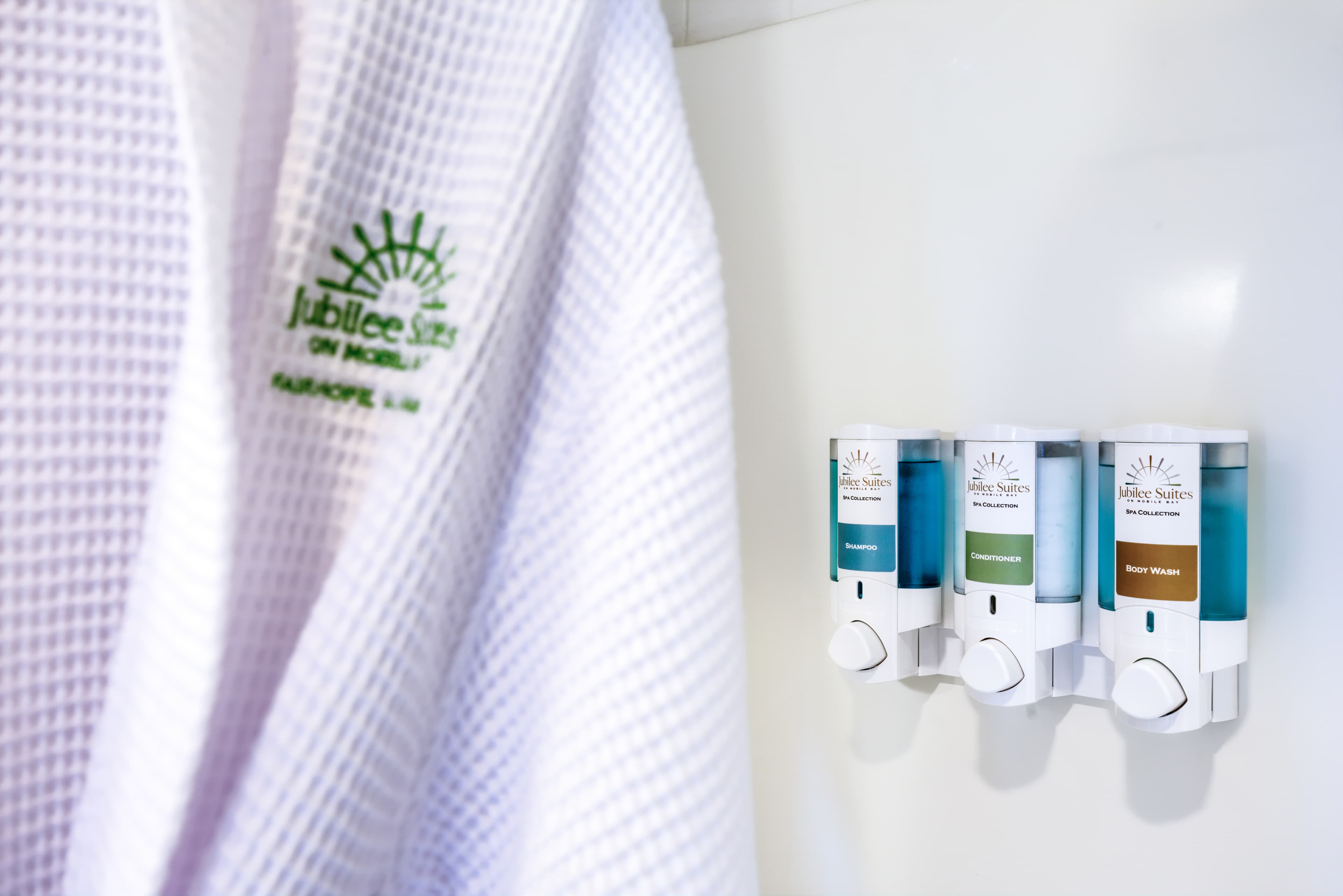 A white waffle-weave bathrobe with embroidered Jubilee Suites logo hangs on a hook in a bathroom. Next to it, a row of dispensers holds various toiletries.