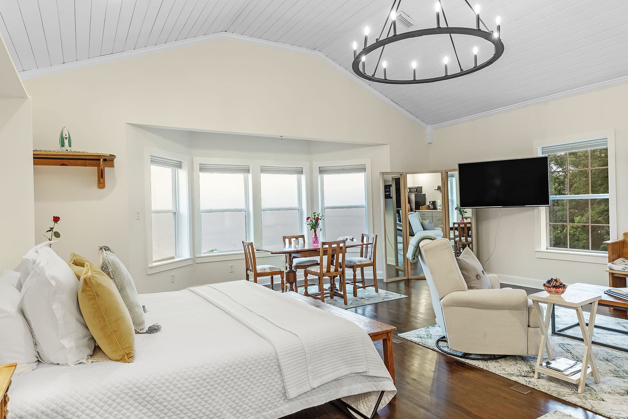 A spacious and inviting bedroom with a queen bed, a sitting area with a recliner and coffee table, and a dining table. The room features a vaulted ceiling with a chandelier, large windows, and hardwood floors.