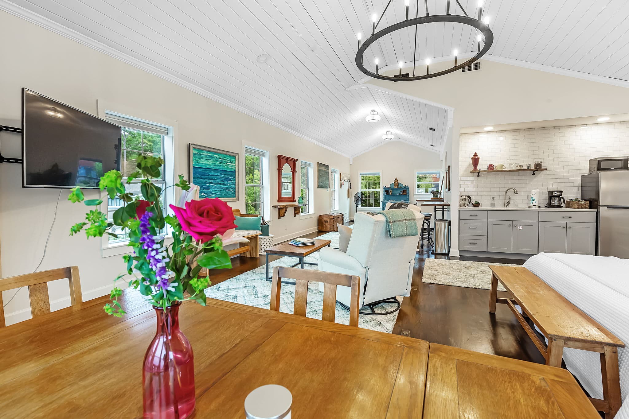 A spacious and inviting open-concept living area with a dining table in the foreground. A vase with colorful flowers adds a touch of vibrancy. Beyond the dining area, a living room with comfortable seating and a TV is visible. A kitchenette is also partially visible in the background.