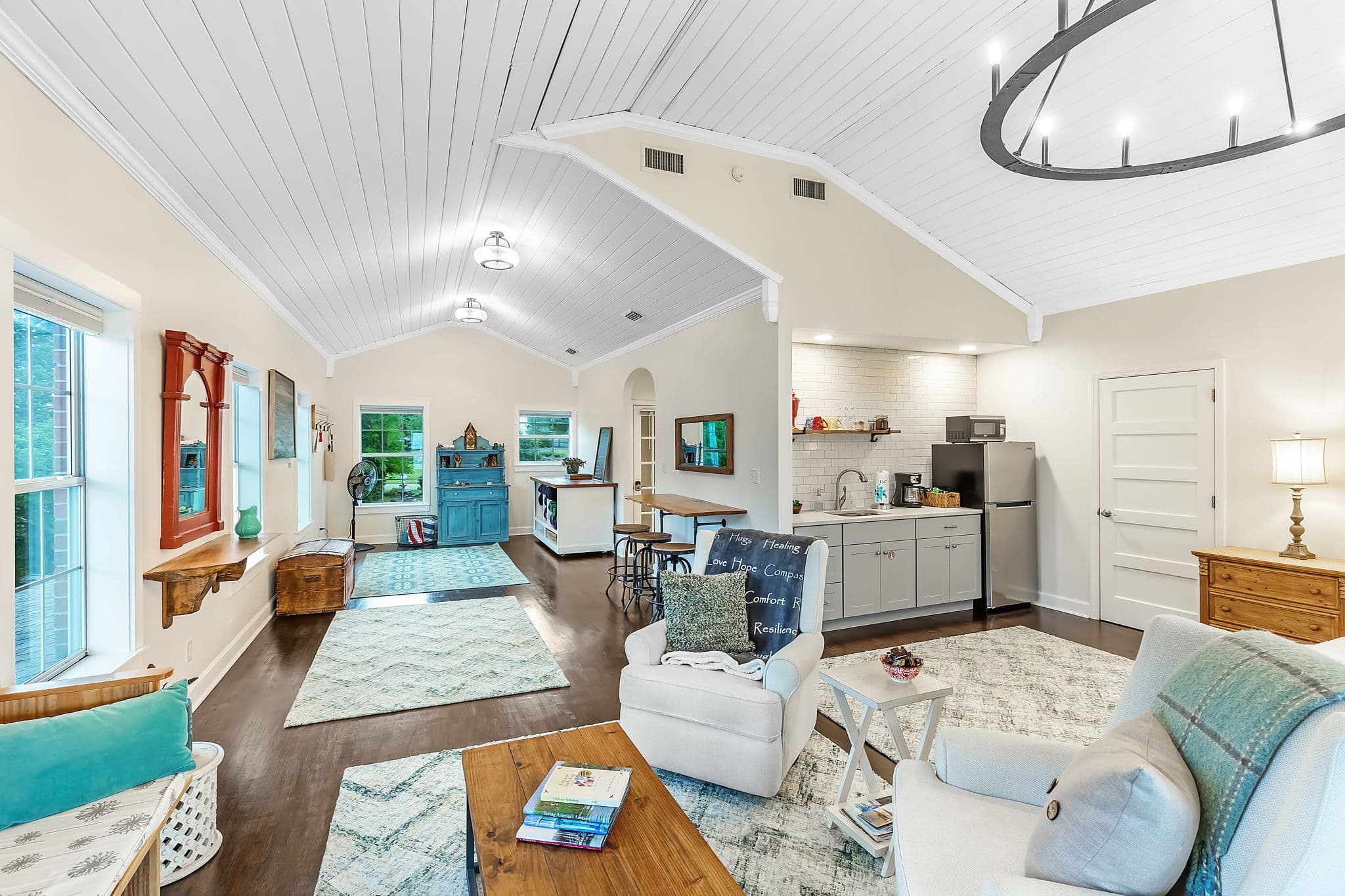 A spacious and open-concept living area with high ceilings and a chandelier. The room features a comfortable seating area with armchairs and a coffee table, a dining table, and a queen-size bed. The space is flooded with natural light and has a warm and inviting atmosphere.