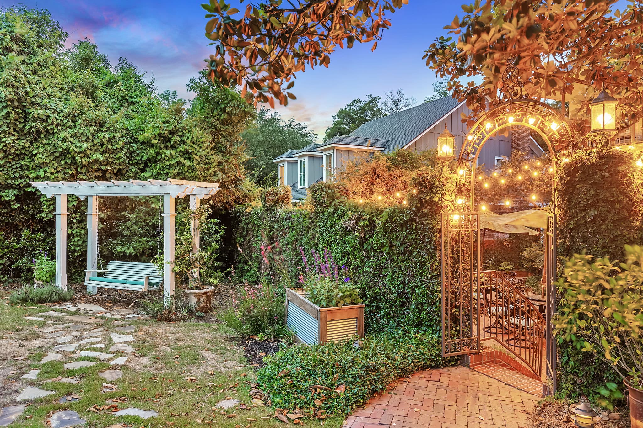 A charming outdoor courtyard with a brick path leading to a gated entrance. String lights illuminate the entrance, creating a warm and inviting atmosphere. A pergola with a swing sits to the side, surrounded by lush greenery.