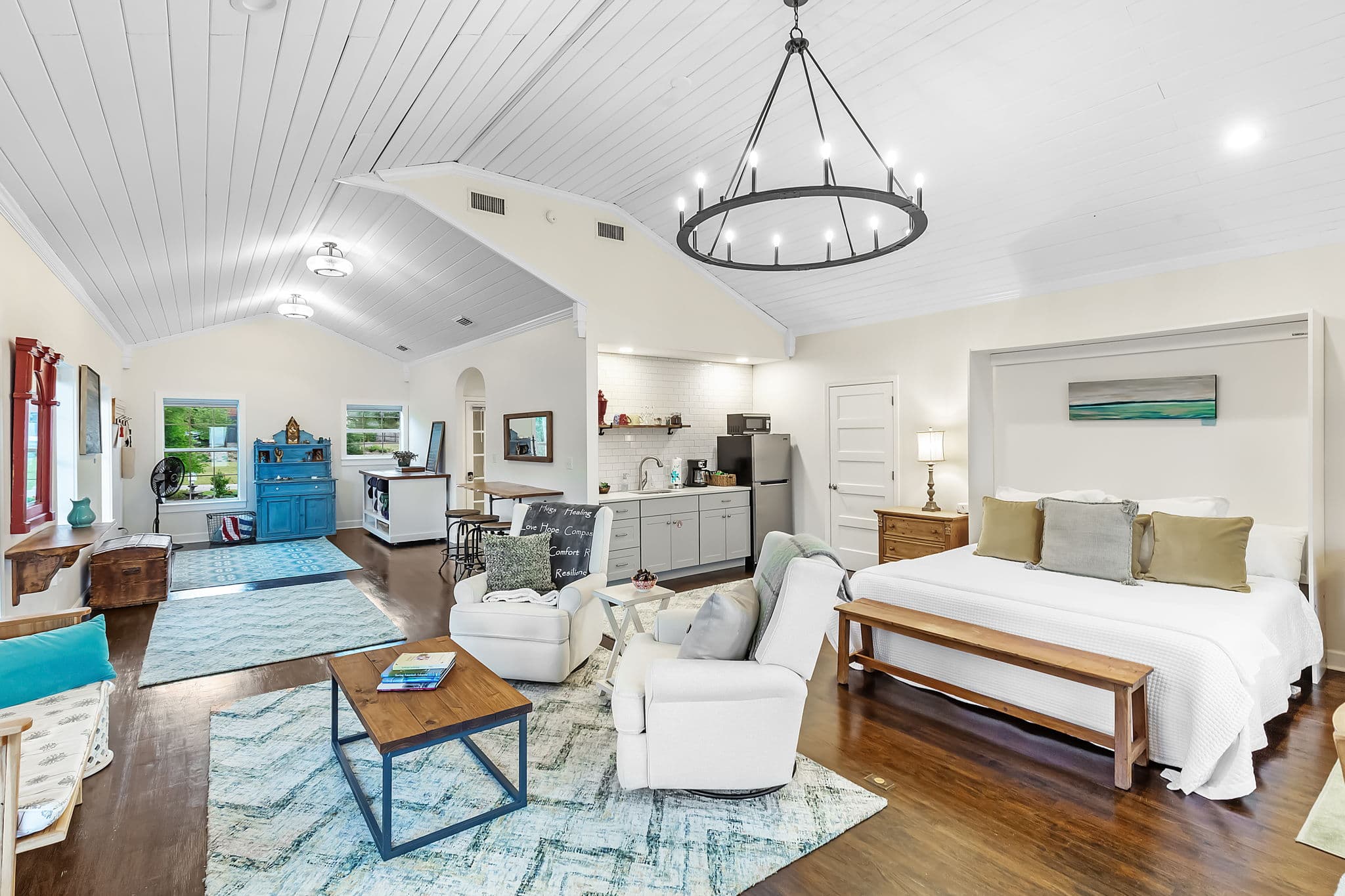 Main living area with a high ceiling and a chandelier. The room features a comfortable seating area with armchairs and a coffee table, a dining table, and a queen bed. The space is flooded with natural light and has a warm and inviting atmosphere.