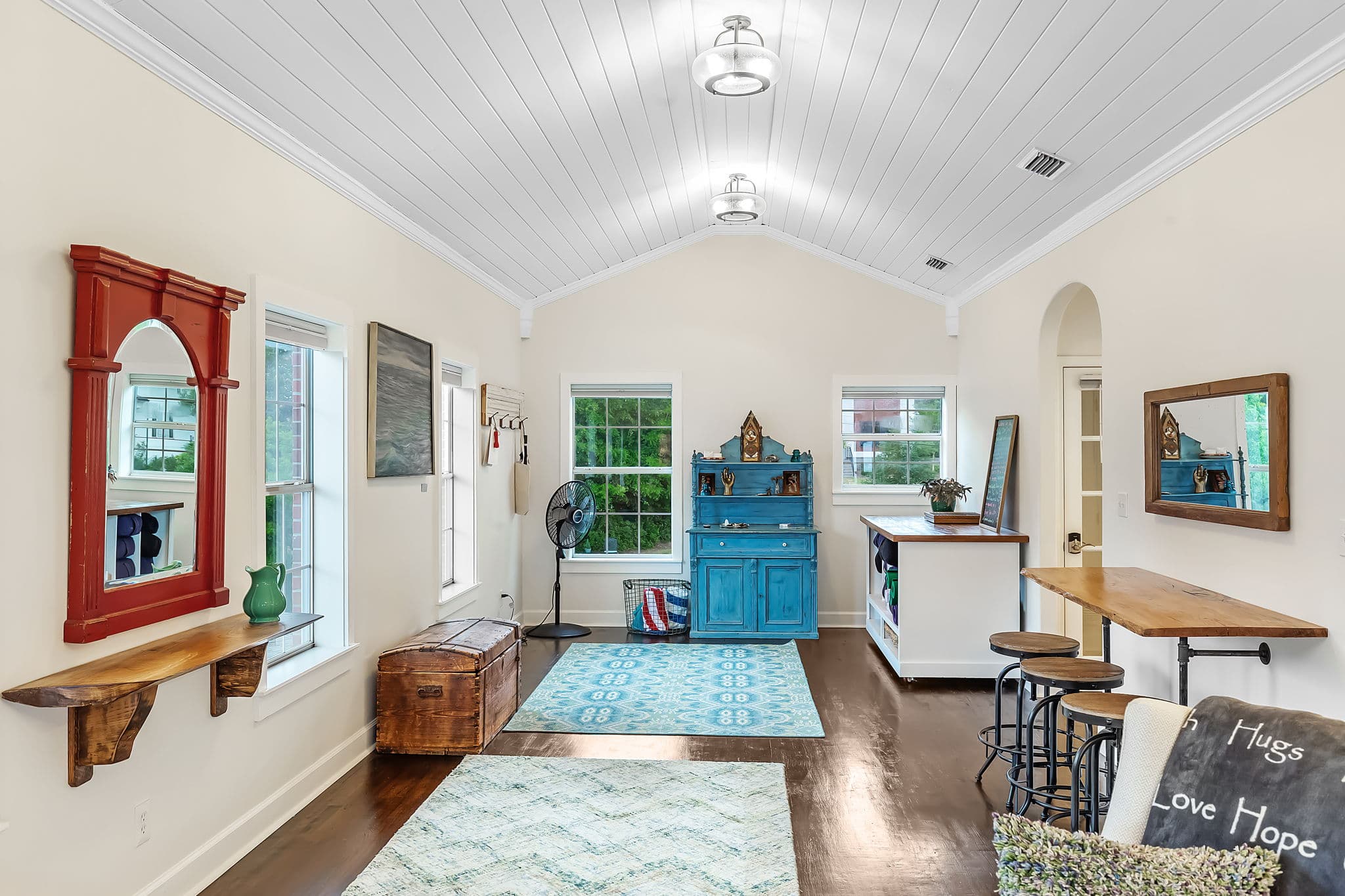 This room features rugs & space for yoga, a vintage dresser, and a wooden table with chairs. There are also several windows that let in natural light.