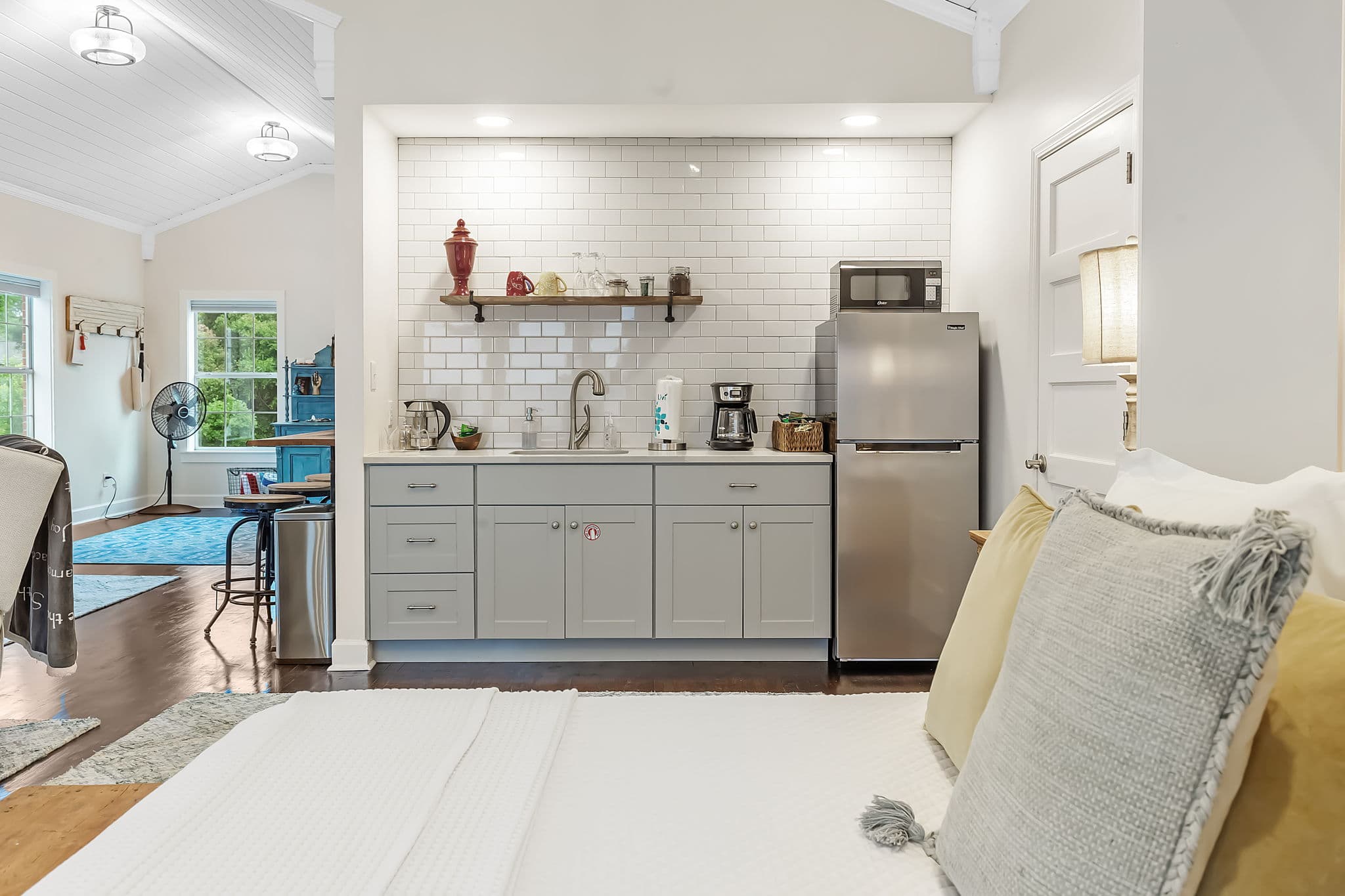 The kitchenette features a small refrigerator, a sink, and cabinets. The bedroom area has a bed with white linens and pillows.