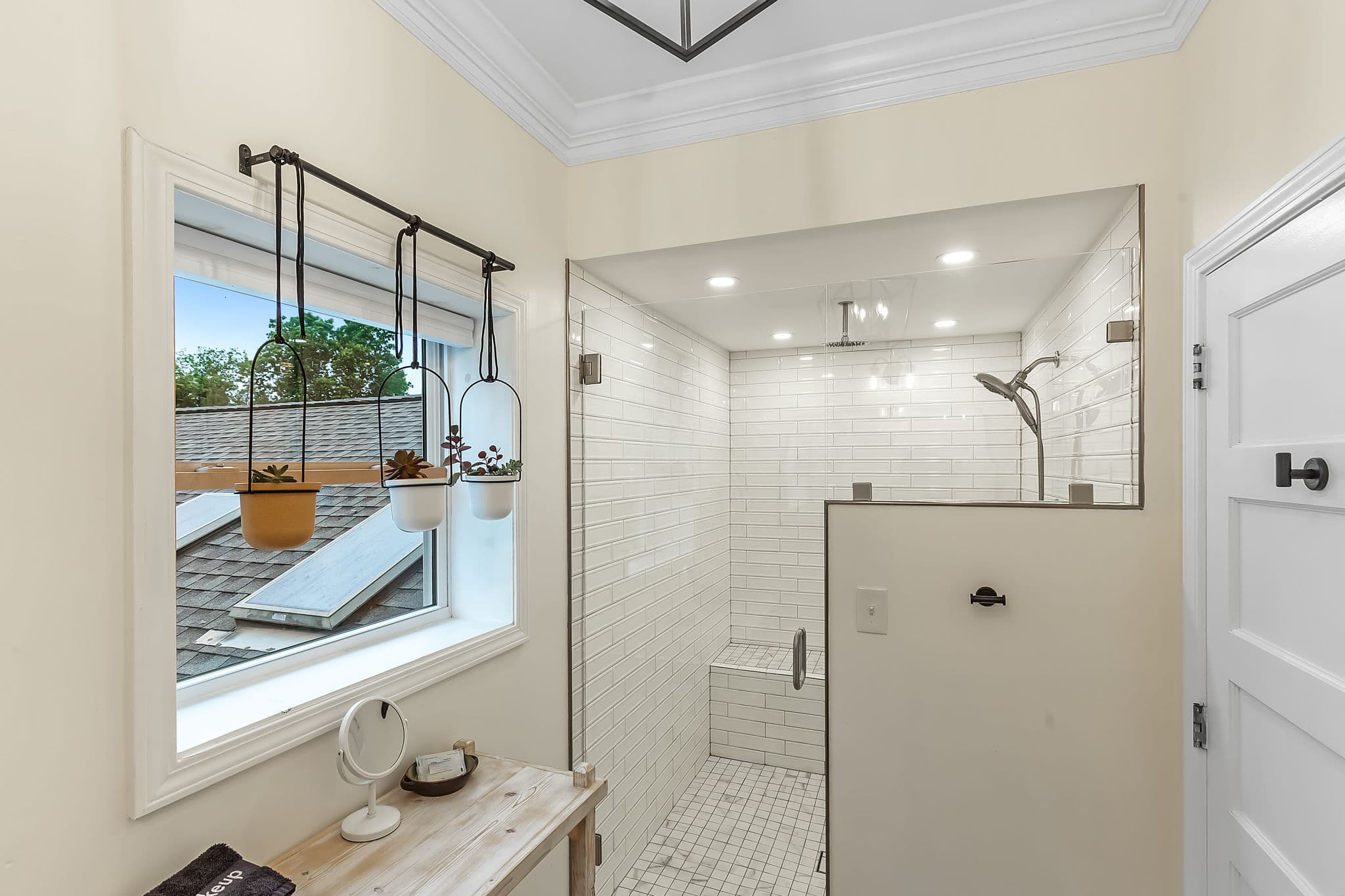 A luxurious bathroom with a large walk-in shower featuring multiple showerheads. The shower has a glass enclosure and is tiled with white subway tiles. A window provides natural light and offers a view of the outdoors. A wooden vanity with a mirror and toiletries completes the space.