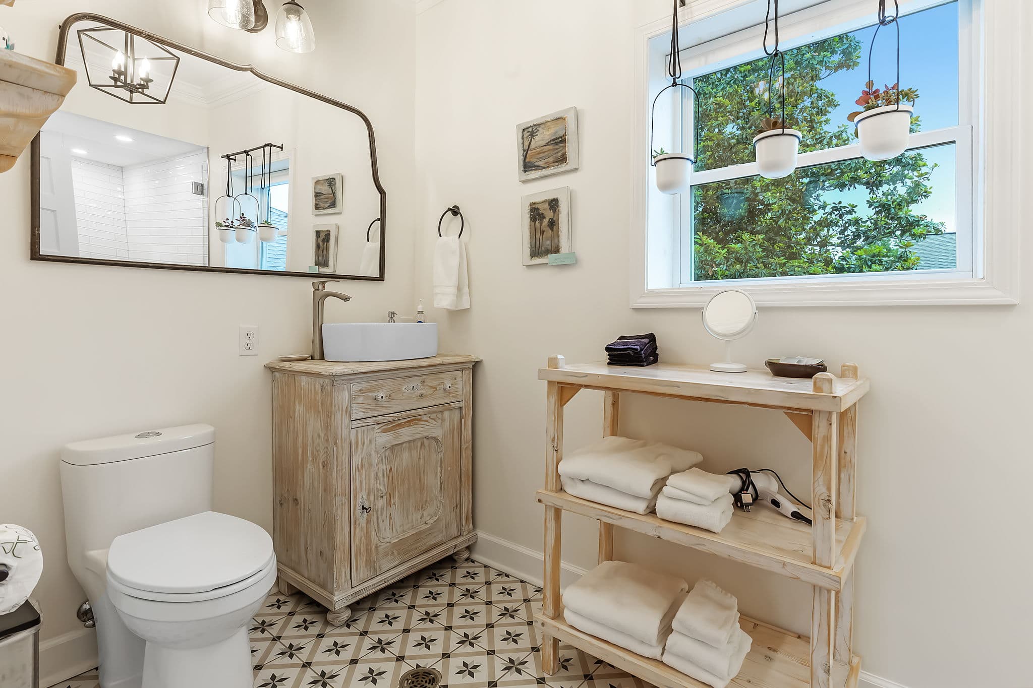 The attached private bathroom is charming with a vintage-style sink and vanity, a toilet, and a shelf holding folded towels. The bathroom has a patterned tile floor and a window with a view of the outdoors.