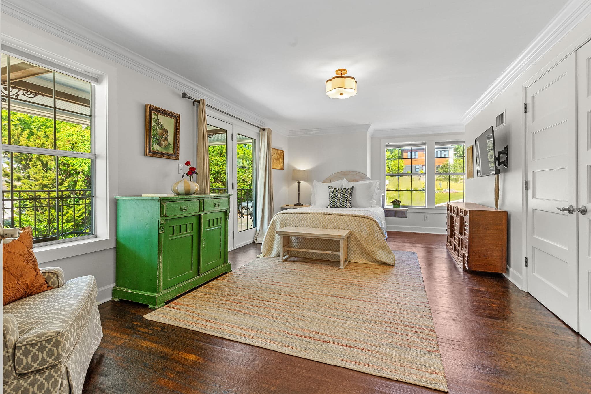A spacious bedroom with large windows overlooking a lush garden. The room features a comfortable queen bed, a green dresser, and a wooden bench.