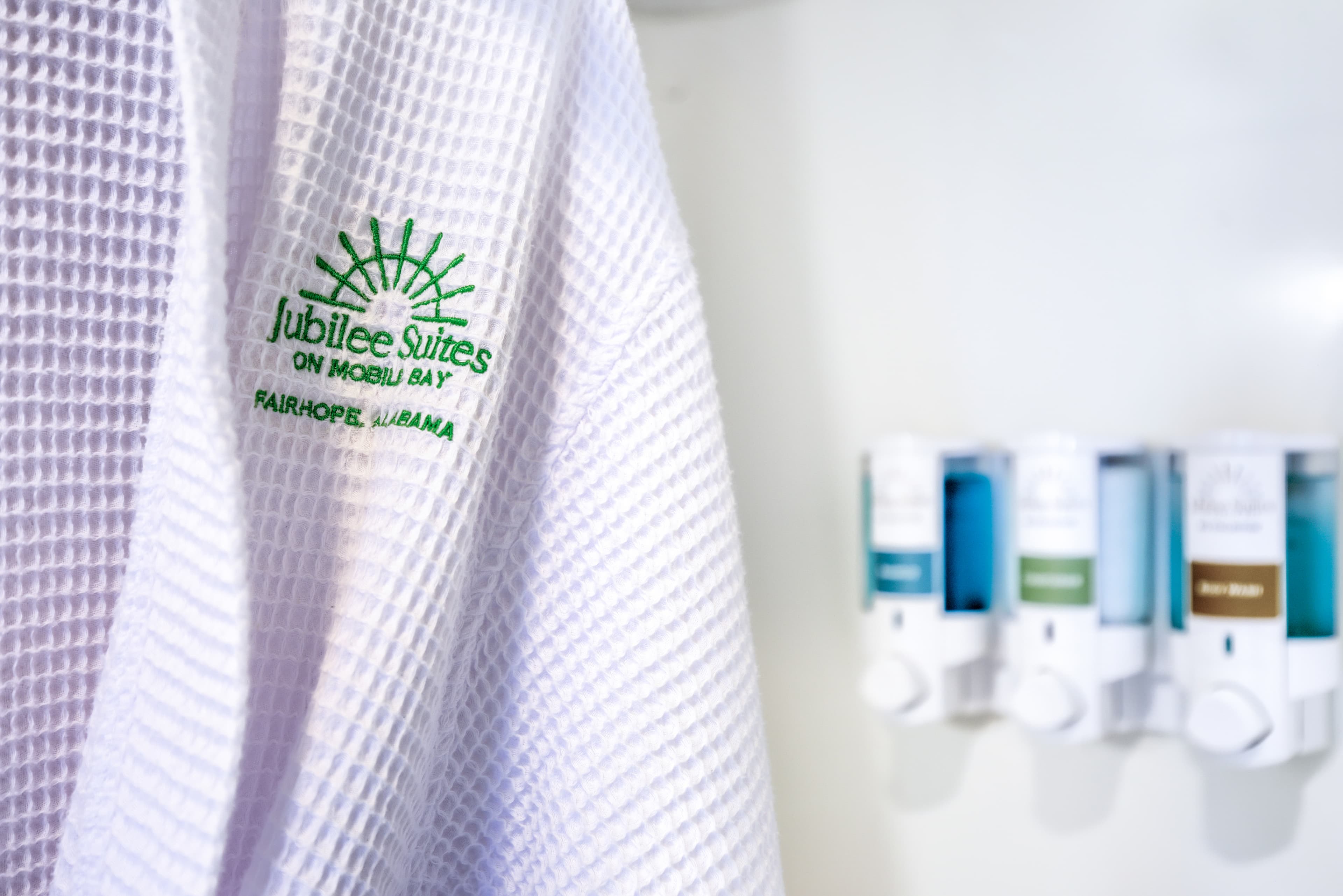 A white bathrobe embroidered in green with the Jubilee Suites logo hangs next to wall mounted soap and shampoo dispensers.
